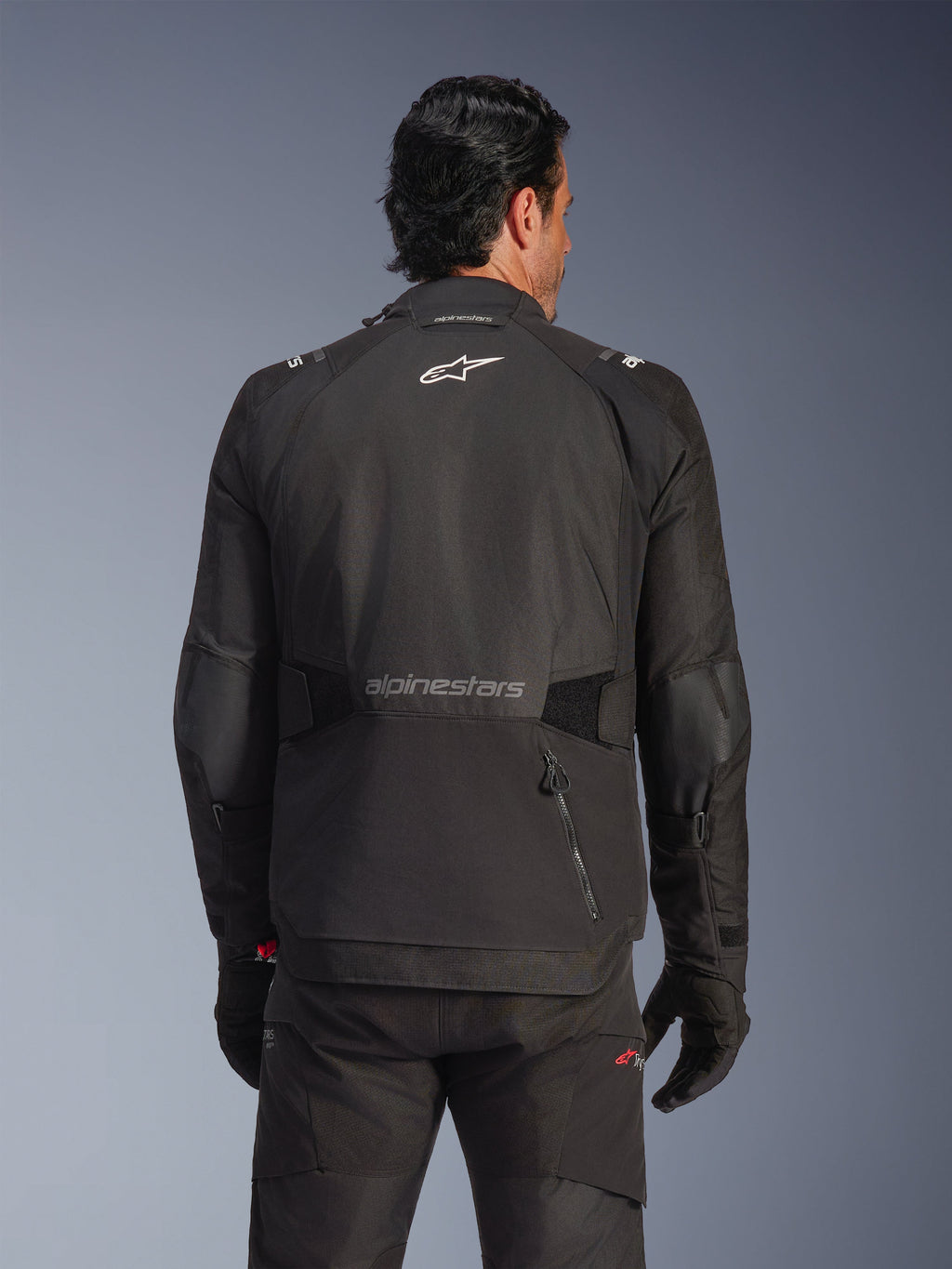 Kurtka Alpinestars Andes V4 Drystar®, czarna, widok z tyłu na modelu, tekstylna kurtka typu adventure z odblaskowymi logotypami i tylną kieszenią zapinaną na zamek, zestawiona z pasującymi czarnymi spodniami i rękawicami