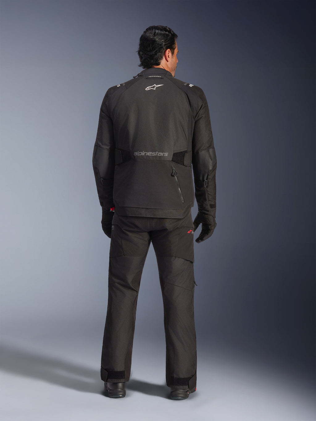 Widok z tyłu na motocyklistę ubranego w kurtkę Alpinestars Andes V4 Drystar® w kolorze czarnym, dopasowane spodnie i rękawice motocyklowe, logo i logotyp Alpinestars na plecach, techniczna odzież adventure na każdą pogodę
