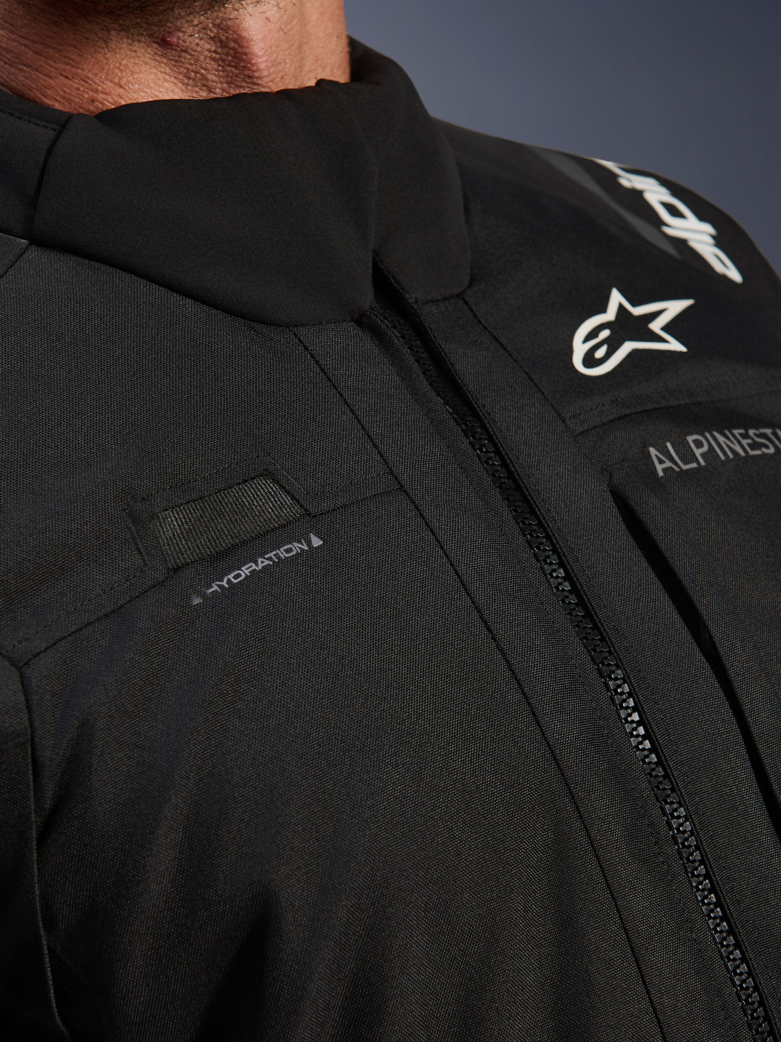 Kurtka Alpinestars Andes V4 Drystar®, czarna, zbliżenie na detale klatki piersiowej z pętlą na wąż nawadniający, wodoodporny przedni zamek VISLON® oraz logo Alpinestars na wytrzymałym materiale tekstylnym