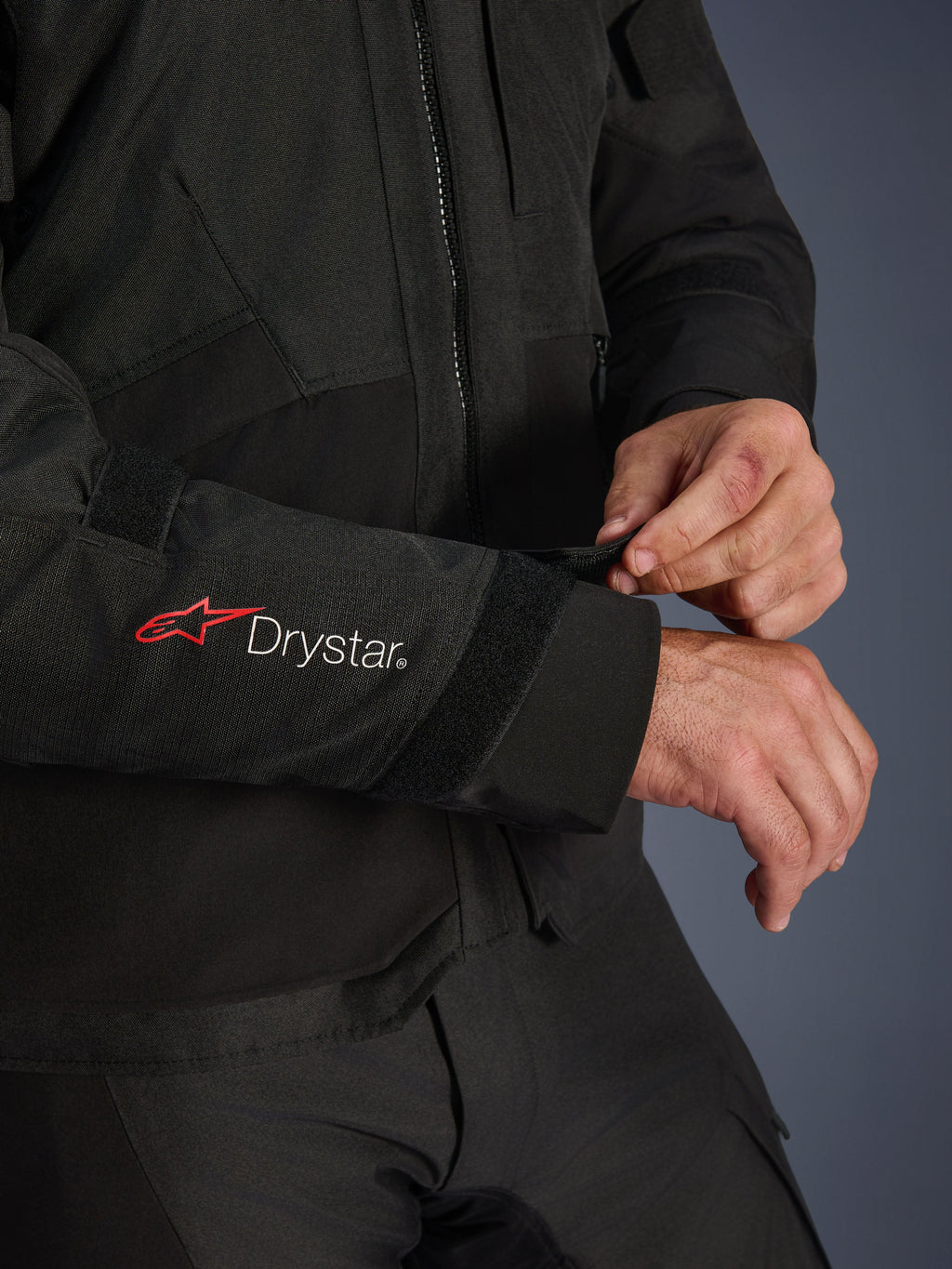 Kurtka Alpinestars Andes V4 Drystar®, czarna tekstylna kurtka motocyklowa, zbliżenie na rękaw z czerwonym logo Alpinestars i białym napisem Drystar®, motocyklista regulujący pasek na rzep przy nadgarstku dla pewnego dopasowania, funkcjonalny detal do jazdy adventure
