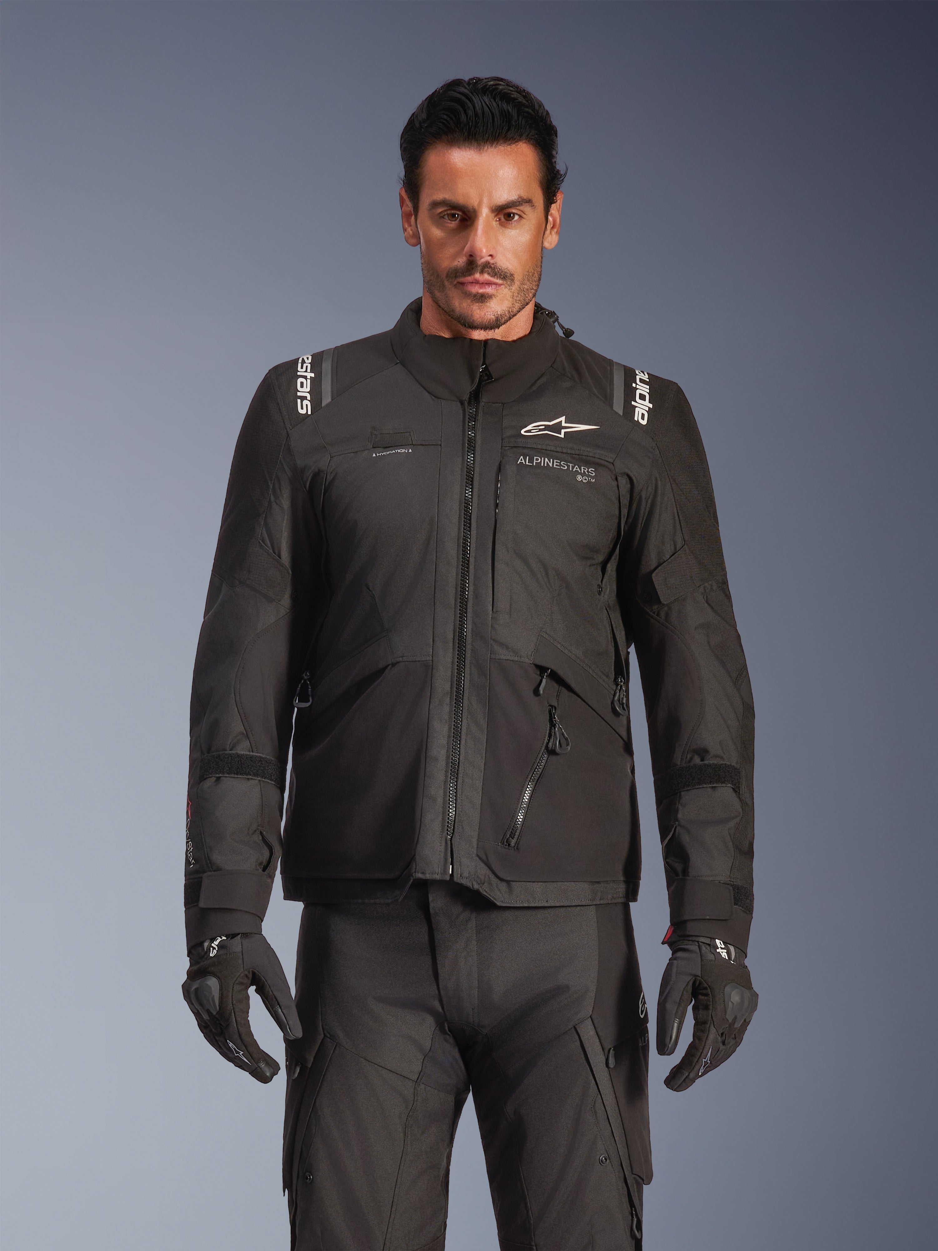 Męski model w kurtce Alpinestars Andes V4 Drystar® w kolorze czarnym, motocyklowa kurtka adventure touring, w zestawie z dopasowanymi czarnymi spodniami i rękawicami, wodoodporne zamki i białe logo Alpinestars na klatce piersiowej i ramionach, zdjęcie studyjne odzieży na każdą pogodę