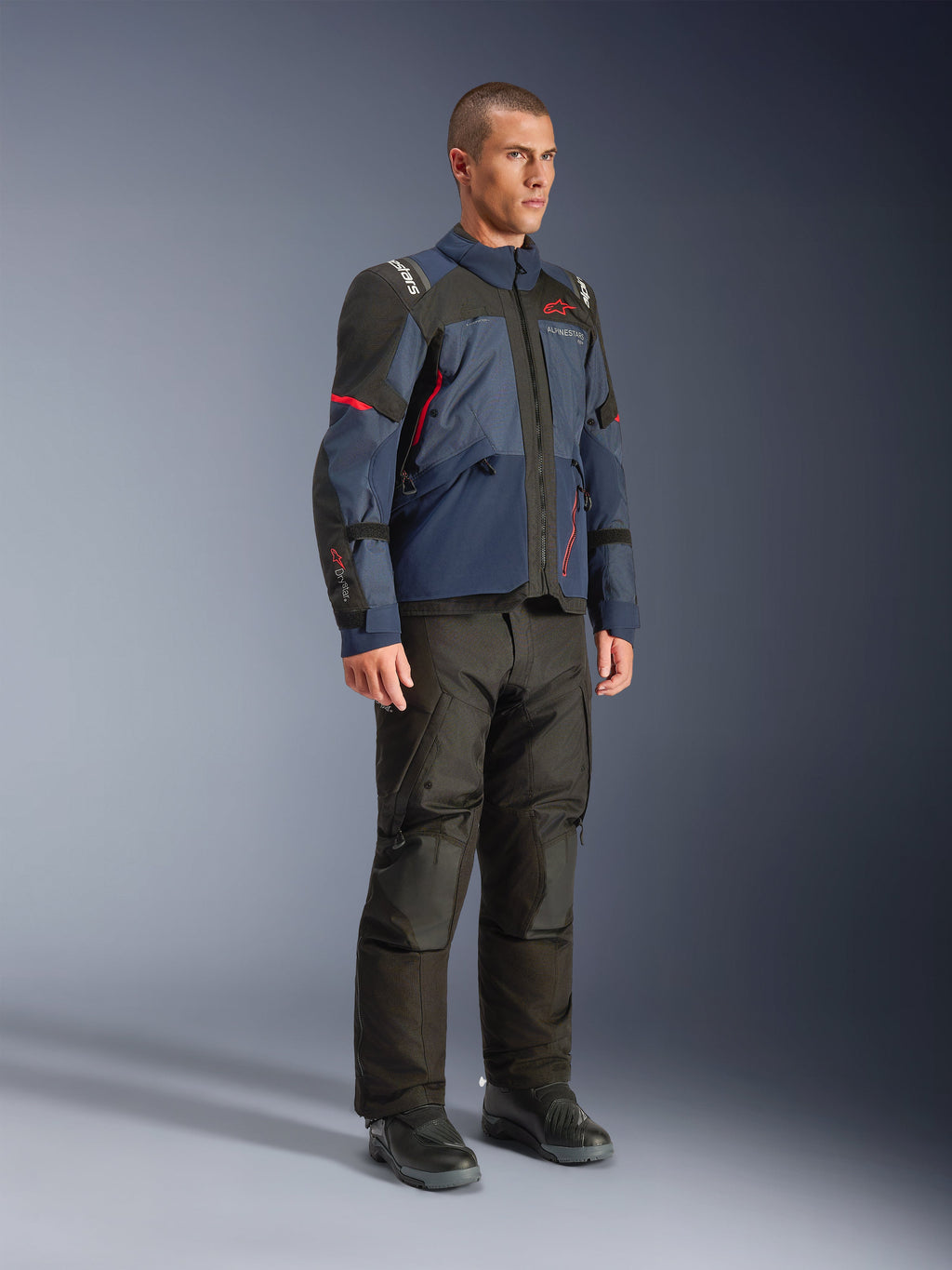Męski model w kurtce Alpinestars Andes V4 Drystar® w kolorystyce Night Navy i Mood Indigo z czarnymi panelami i czerwonymi akcentami, zestawionej z czarnymi spodniami i butami motocyklowymi, techniczna odzież adventure touring w studio.