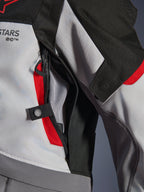 Close-up detail of the Alpinestars Andes V4 Drystar® Jacket in Dark Gray, Ice Gray, and Bright Red, highlighting a zippered chest ventilation port with mesh lining and a snap button adjustment strap.