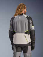Kurtka Alpinestars Andes V4 Drystar®, widok od tyłu przygodowej kurtki motocyklowej w kolorach ciemnoszarym, jasnoszarym i czarnym z jaskrawożółtymi akcentami fluo, wzmocnione panele tekstylne i logo Alpinestars, noszona z pasującymi spodniami i czarnymi rękawicami