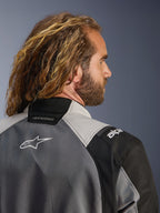 Zbliżenie na profil z tyłu mężczyzny z długimi, falowanymi włosami w kurtce Alpinestars Andes V4 Drystar® w kolorze ciemnoszarym i jasnoszarym, z tekstylnymi panelami i białym logo Alpinestars na plecach, motocyklowa odzież adventure