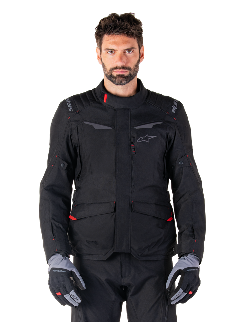 Alpinestars ST-1 Waterproof Jacket, Black motorcycle adventure touring jacket, worn by a male model with matching black pants and grey/black gloves, featuring minimalistic styling, Alpinestars branding on the shoulders, large front cargo pockets with red details, and ergonomic construction for all-weather road touring.