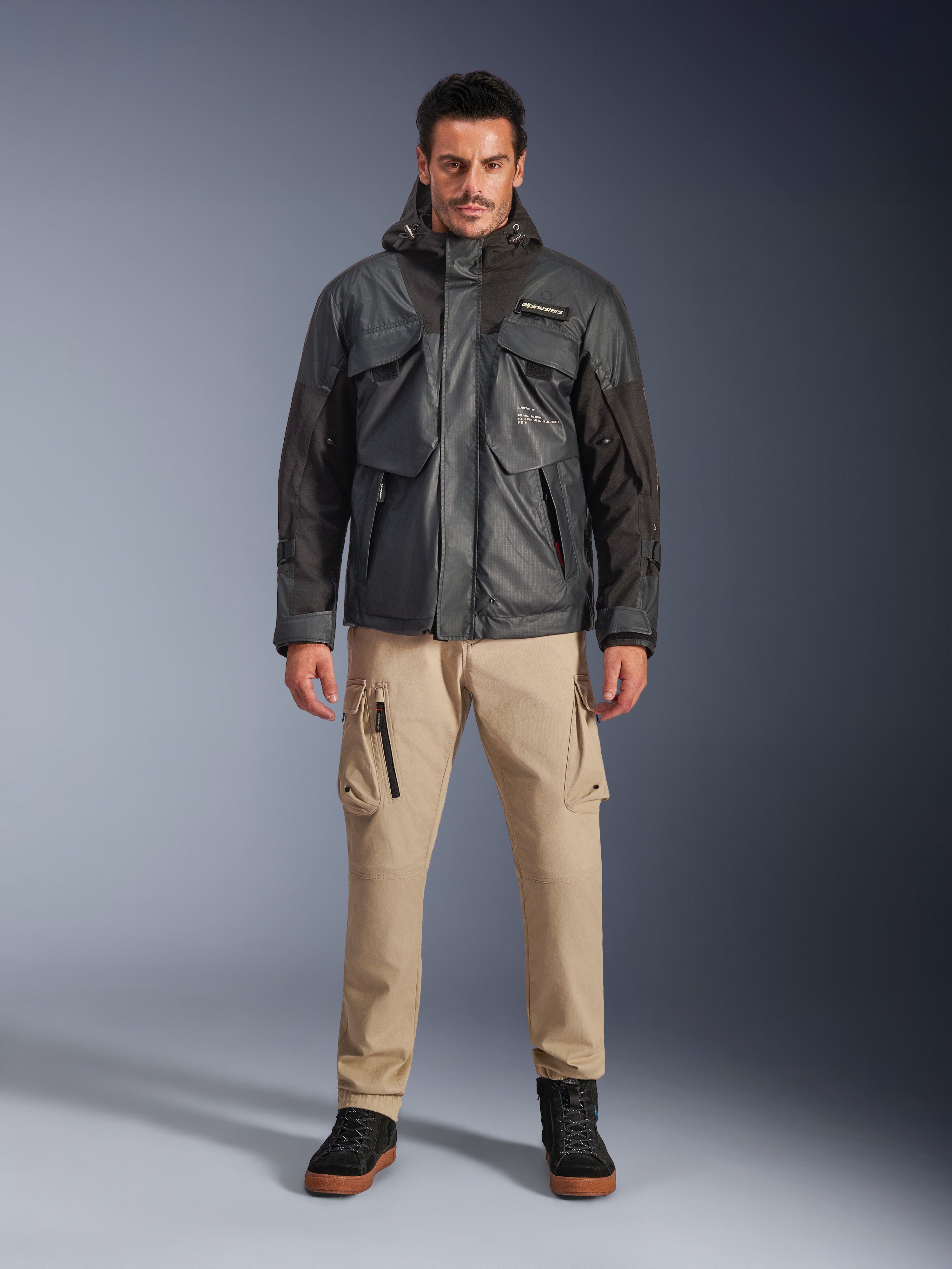 Męski model w kurtce Alpinestars Mospher Waterproof w kolorze czarnym, nowoczesna miejska odzież motocyklowa, taktyczny design użytkowy z wieloma funkcjonalnymi kieszeniami i kapturem, beżowe spodnie cargo i czarne buty, portret studyjny całej sylwetki.