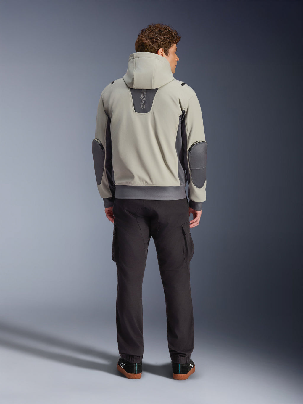 Alpinestars Moblast Waterproof Jacket, kolor Iron Gray Dark Sage, widok z tyłu na motocyklistę w kurtce motocyklowej z kapturem, jasnoszara i ciemnoszałwiowa kolorystyka, pionowe logo „astars” w górnej części pleców, ciemnoszare łaty na łokciach, stylizacja z ciemnymi spodniami cargo dla nowoczesnego miejskiego wyglądu.
