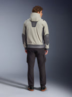 Alpinestars Moblast Waterproof Jacket, kolor Iron Gray Dark Sage, widok z tyłu na motocyklistę w kurtce motocyklowej z kapturem, jasnoszara i ciemnoszałwiowa kolorystyka, pionowe logo „astars” w górnej części pleców, ciemnoszare łaty na łokciach, stylizacja z ciemnymi spodniami cargo dla nowoczesnego miejskiego wyglądu.