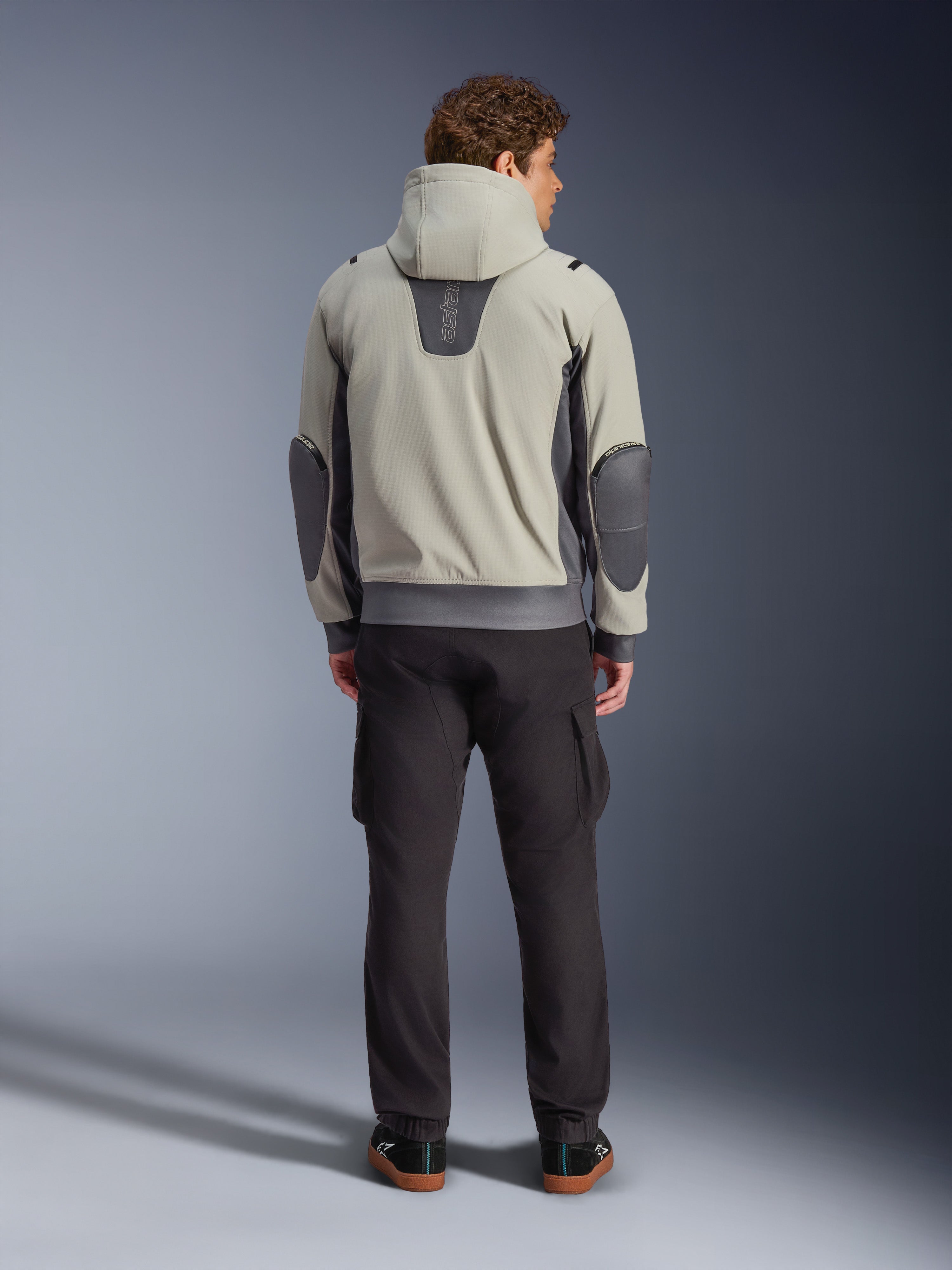 Alpinestars Moblast Waterproof Jacket, kolor Iron Gray Dark Sage, widok z tyłu na motocyklistę w kurtce motocyklowej z kapturem, jasnoszara i ciemnoszałwiowa kolorystyka, pionowe logo „astars” w górnej części pleców, ciemnoszare łaty na łokciach, stylizacja z ciemnymi spodniami cargo dla nowoczesnego miejskiego wyglądu.