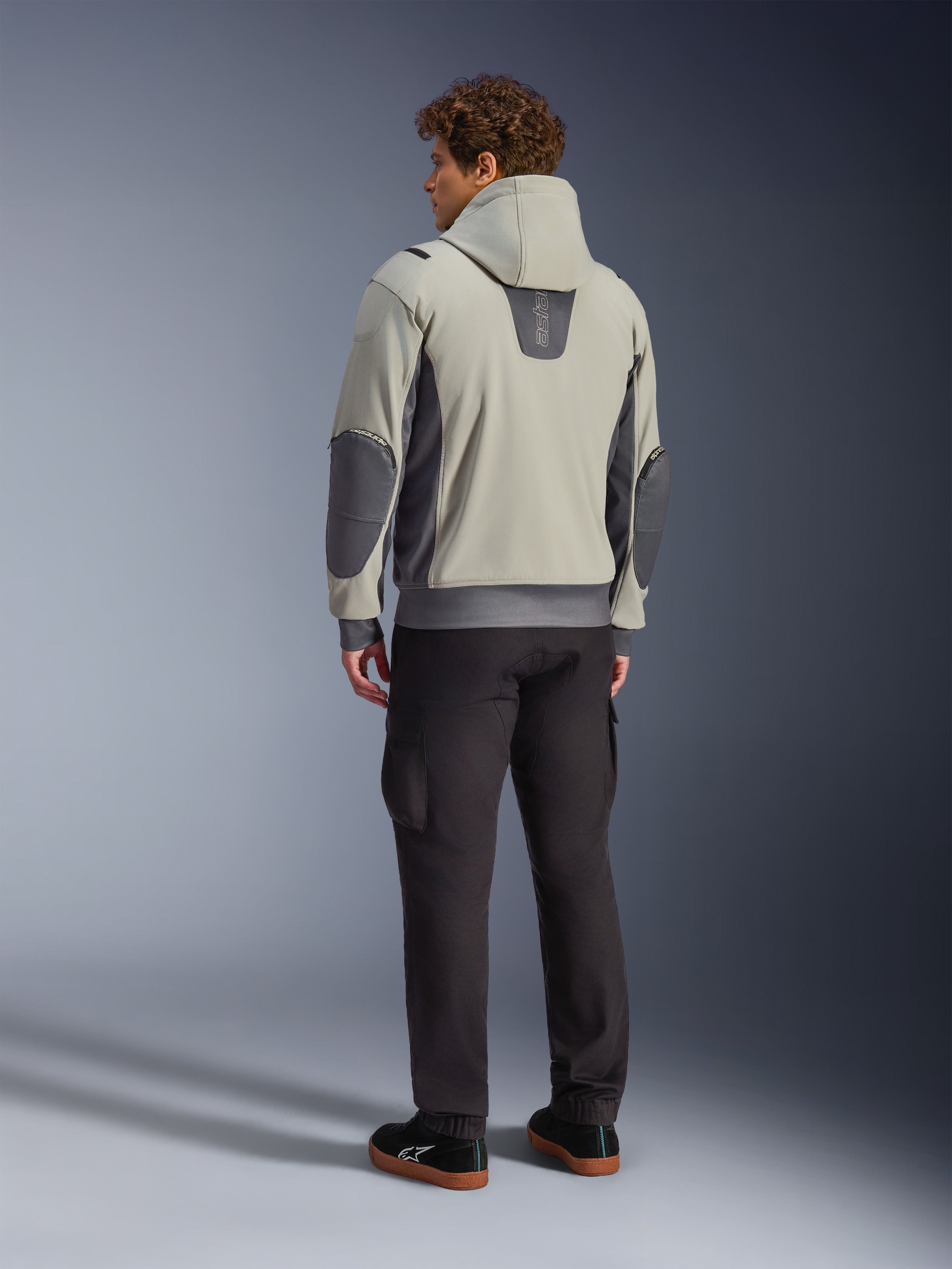 Męski model widoczny od tyłu w kurtce Alpinestars Moblast Waterproof Jacket w kolorze Iron Gray Dark Sage, miejska kurtka motocyklowa z kapturem, ciemnoszarymi panelami bocznymi i wzmocnionymi łatami na łokciach, w zestawieniu z ciemnymi spodniami motocyklowymi i czarnymi tenisówkami, nowoczesna minimalistyczna estetyka do miejskich dojazdów
