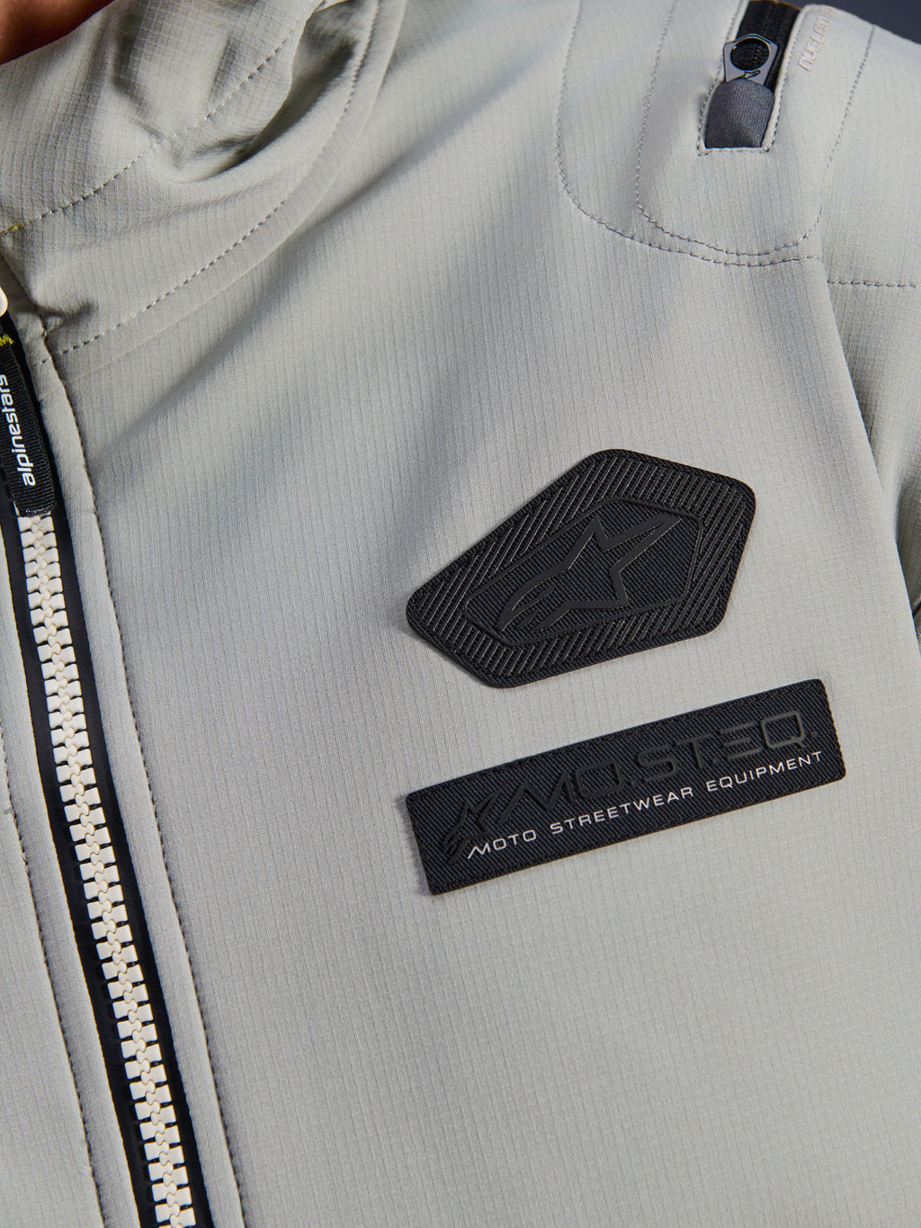 Kurtka Alpinestars Moblast Waterproof, Iron Gray, zbliżenie na naszywki na klatce piersiowej z logo Alpinestars i brandingiem MO.ST.EQ. Moto Streetwear Equipment, techniczna tkanina ripstop, solidny zamek błyskawiczny oraz detal kieszeni na ramieniu.