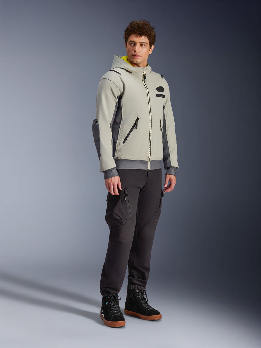 Alpinestars Moblast Waterproof Jacket, Iron Gray i Dark Sage, miejska kurtka motocyklowa o fasonie bluzy z kapturem, prezentowana przez modela w ciemnych spodniach i czarnych butach, minimalistyczna estetyka cyber-tech z żółtym wnętrzem kaptura i wodoodpornymi zamkami