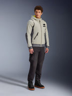 Alpinestars Moblast Waterproof Jacket, Iron Gray i Dark Sage, miejska kurtka motocyklowa o fasonie bluzy z kapturem, prezentowana przez modela w ciemnych spodniach i czarnych butach, minimalistyczna estetyka cyber-tech z żółtym wnętrzem kaptura i wodoodpornymi zamkami