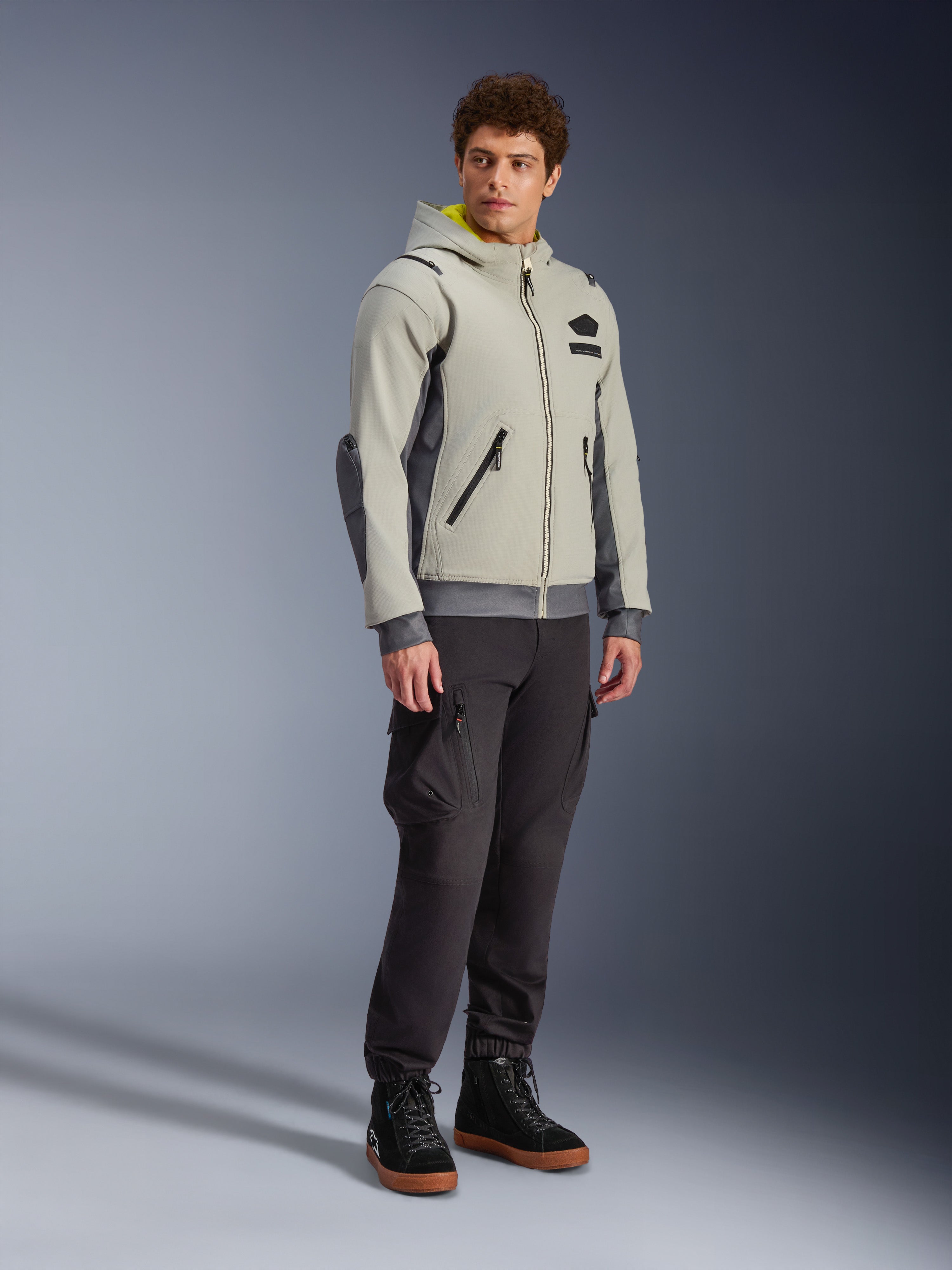 Alpinestars Moblast Waterproof Jacket, Iron Gray i Dark Sage, miejska kurtka motocyklowa o fasonie bluzy z kapturem, prezentowana przez modela w ciemnych spodniach i czarnych butach, minimalistyczna estetyka cyber-tech z żółtym wnętrzem kaptura i wodoodpornymi zamkami