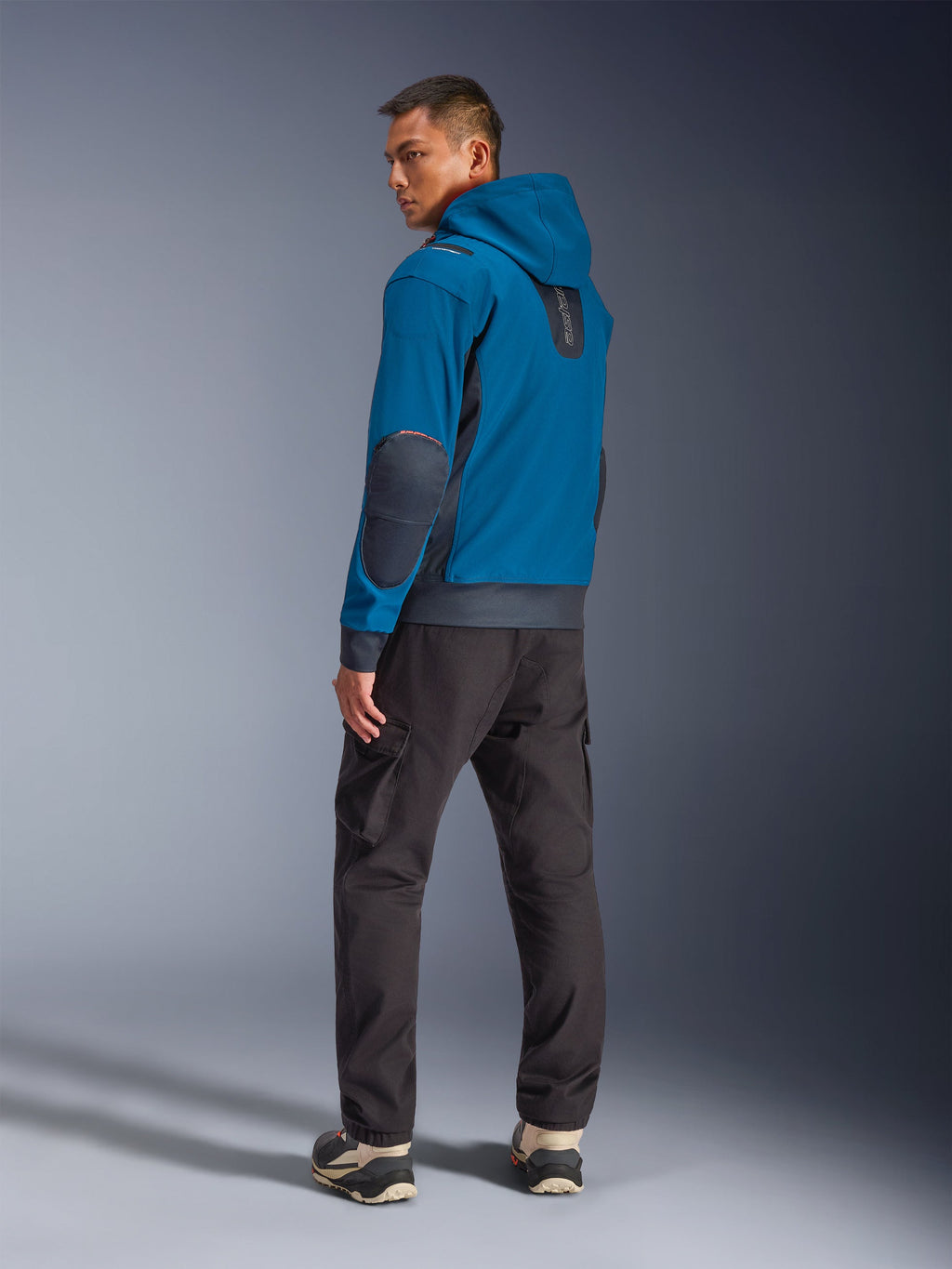 Alpinestars Moblast Waterproof Jacket w kolorze Dark Petrol i Dark Navy, widok z tyłu na modelu prezentujący fason z kapturem, granatowe łaty na łokciach i boczne wstawki, zestawiona z ciemnymi spodniami cargo i technicznymi sneakersami, nowoczesna miejska odzież motocyklowa