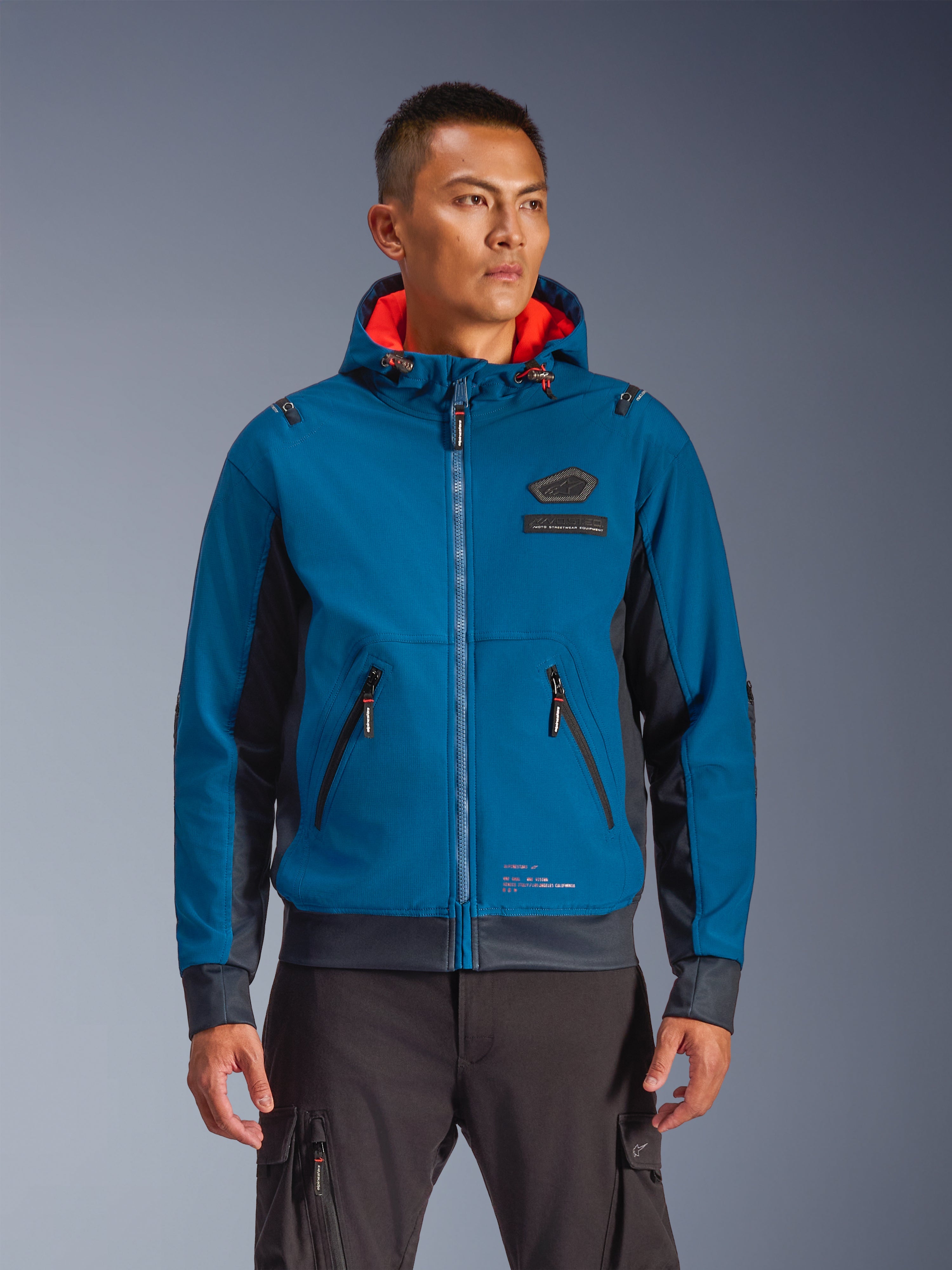 Alpinestars Moblast Waterproof Jacket prezentowana przez modela, niebieska i ciemnogranatowa miejska kurtka motocyklowa, kaptur z czerwoną podszewką, czarne zamki i naszywka na klatce piersiowej, odzież motocyklowa inspirowana stylem streetwear