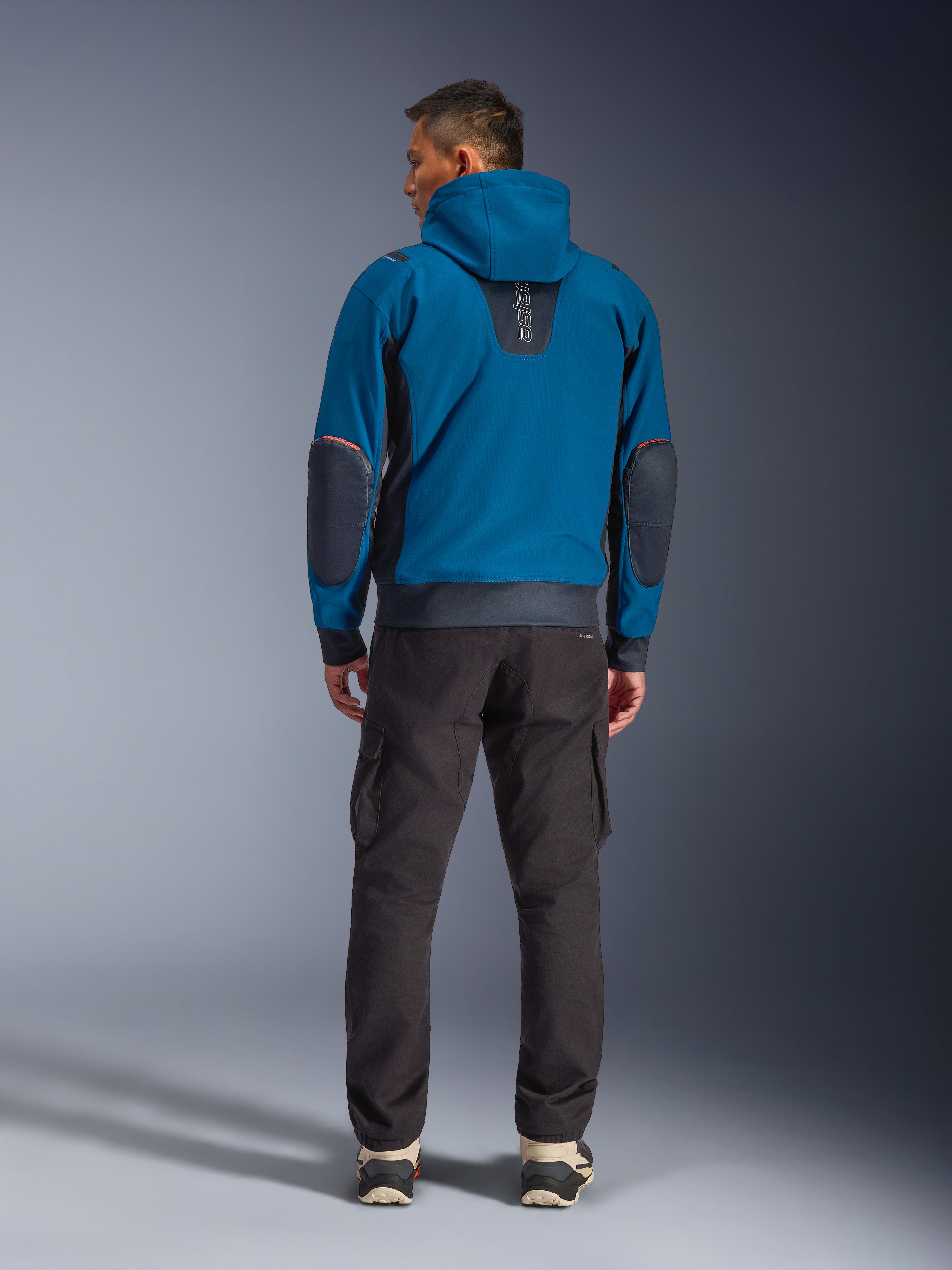 Alpinestars Moblast Waterproof Jacket, miejska kurtka motocyklowa, kolory Dark Petrol i Dark Navy Blue, widok z tyłu ukazujący kaptur i wzmocnione łaty na łokciach, noszona z ciemnymi spodniami cargo, stylowa odzież motocyklowa na każdą pogodę