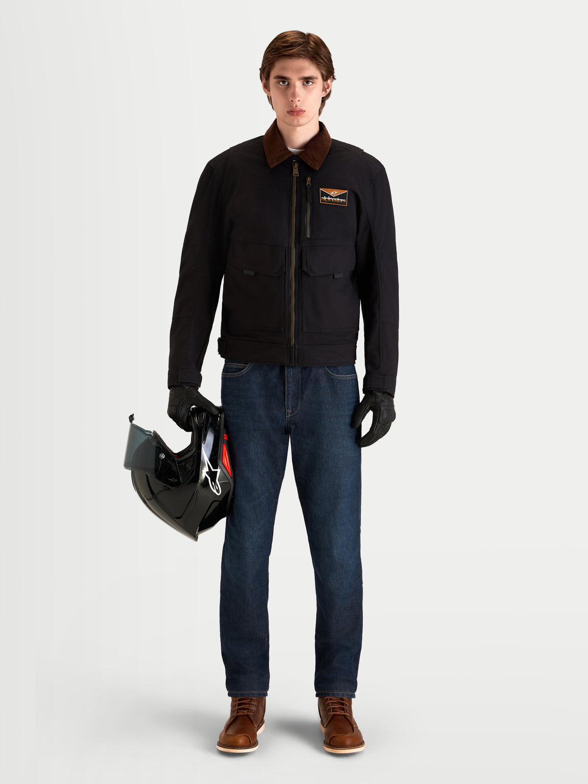 Alpinestars Mustang Wp Kurtka, męska współczesna miejska kurtka motocyklowa w kolorze ciemnego antracytu i ciemnego brązu, widok z przodu na modelu w ochronnym sprzęcie motocyklowym trzymającym kask, prezentująca inspirowaną odzieżą roboczą konstrukcję z elastycznego twillu oraz sztruksowy kołnierz