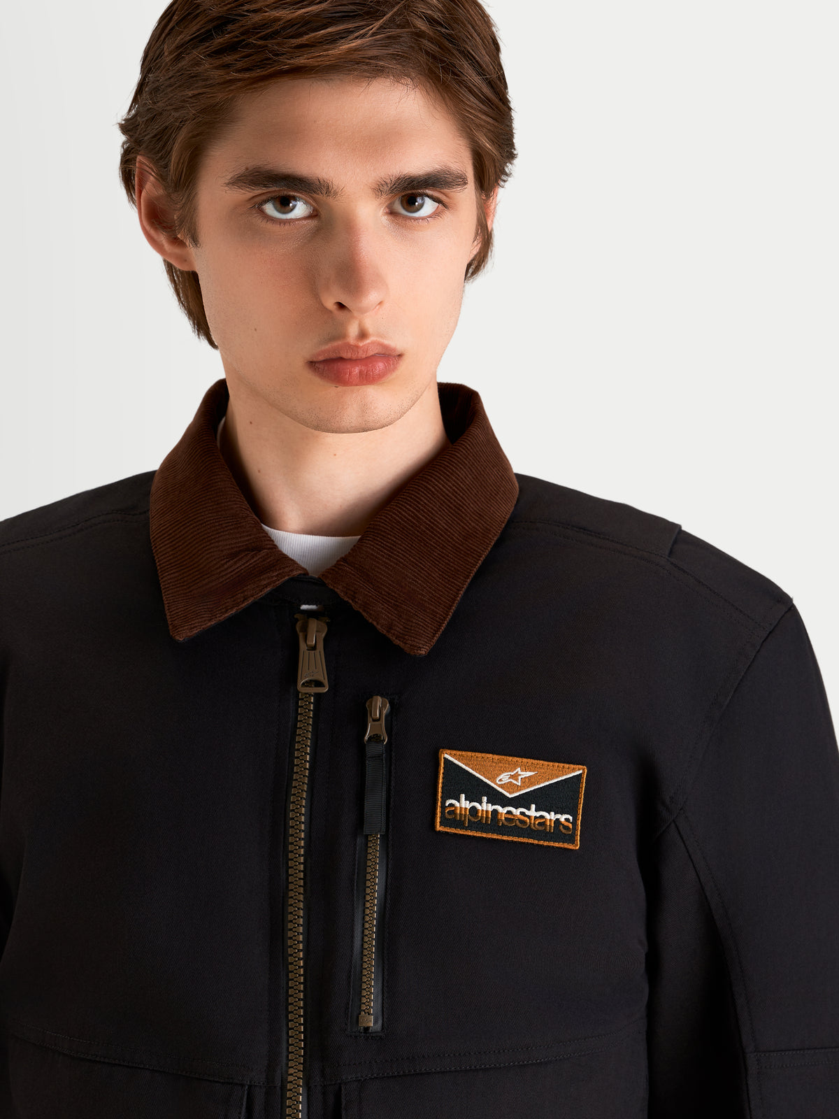 Alpinestars Mustang Wp Kurtka, męska miejska kurtka motocyklowa w kolorze ciemnoszarym i ciemnobrązowym, zbliżenie przedstawiające kołnierz ze sztruksu inspirowany odzieżą roboczą, przedni zamek błyskawiczny i naszywkę z logo na klatce piersiowej noszoną przez modela
