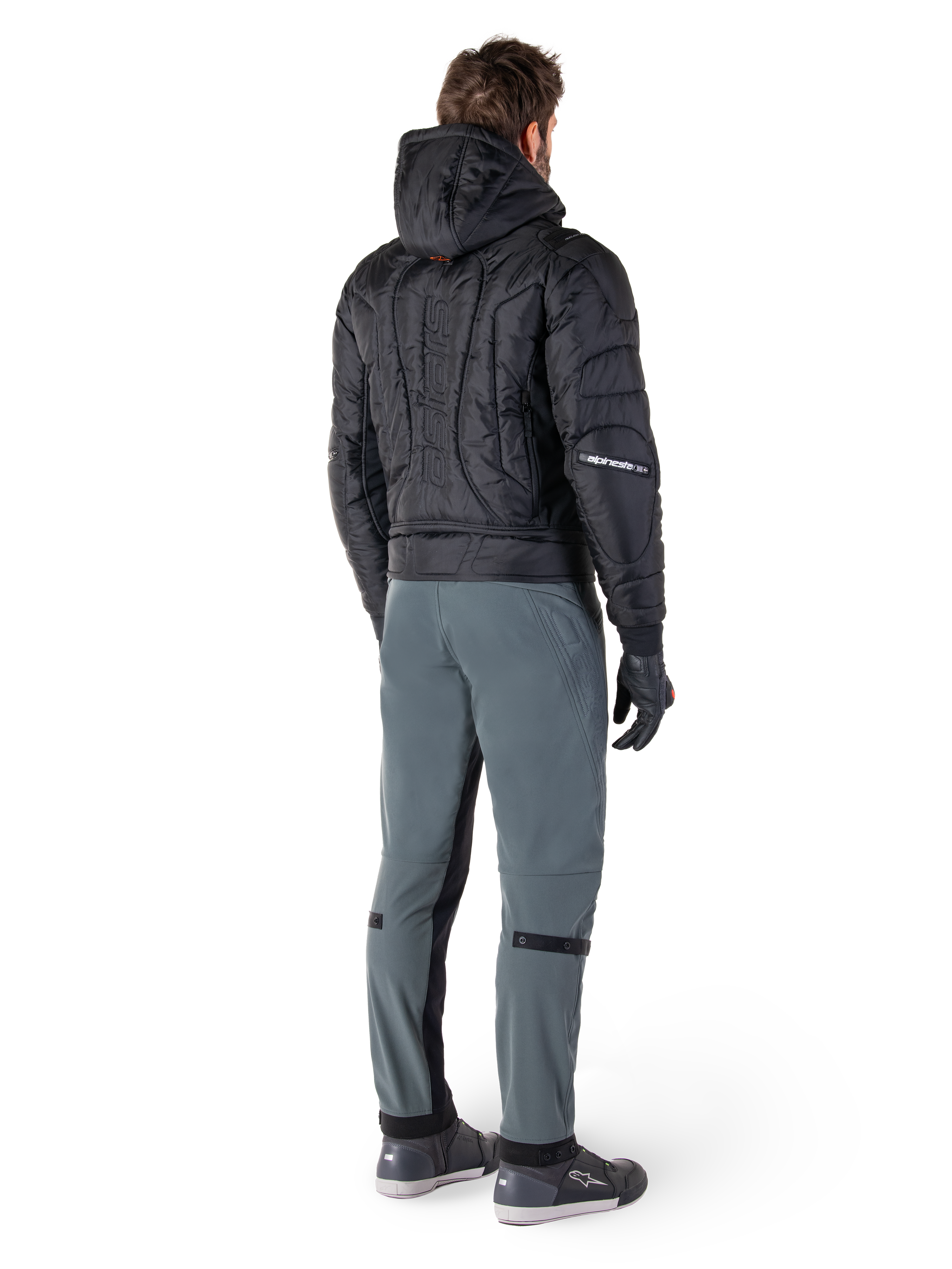 Alpinestars Mohobbs Waterproof Jacket, czarna, widok z tyłu na pikowaną kurtkę bomber z wytłoczonym pionowym napisem na plecach, zestawiona z szarymi spodniami motocyklowymi i czarnymi rękawicami, miejska odzież motocyklowa inspirowana stylem streetwear