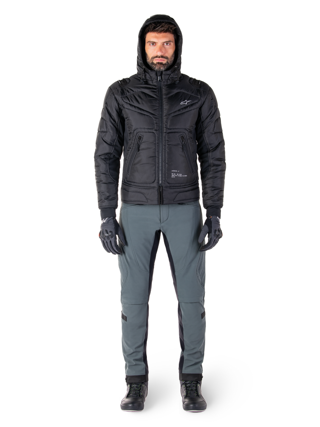 Kurtka Alpinestars Mohobbs Waterproof w kolorze czarnym, męski model w pikowanej kurtce typu bomber z kapturem, zestawiona z szarymi spodniami motocyklowymi, czarnymi rękawicami i butami, miejska współczesna odzież motocyklowa