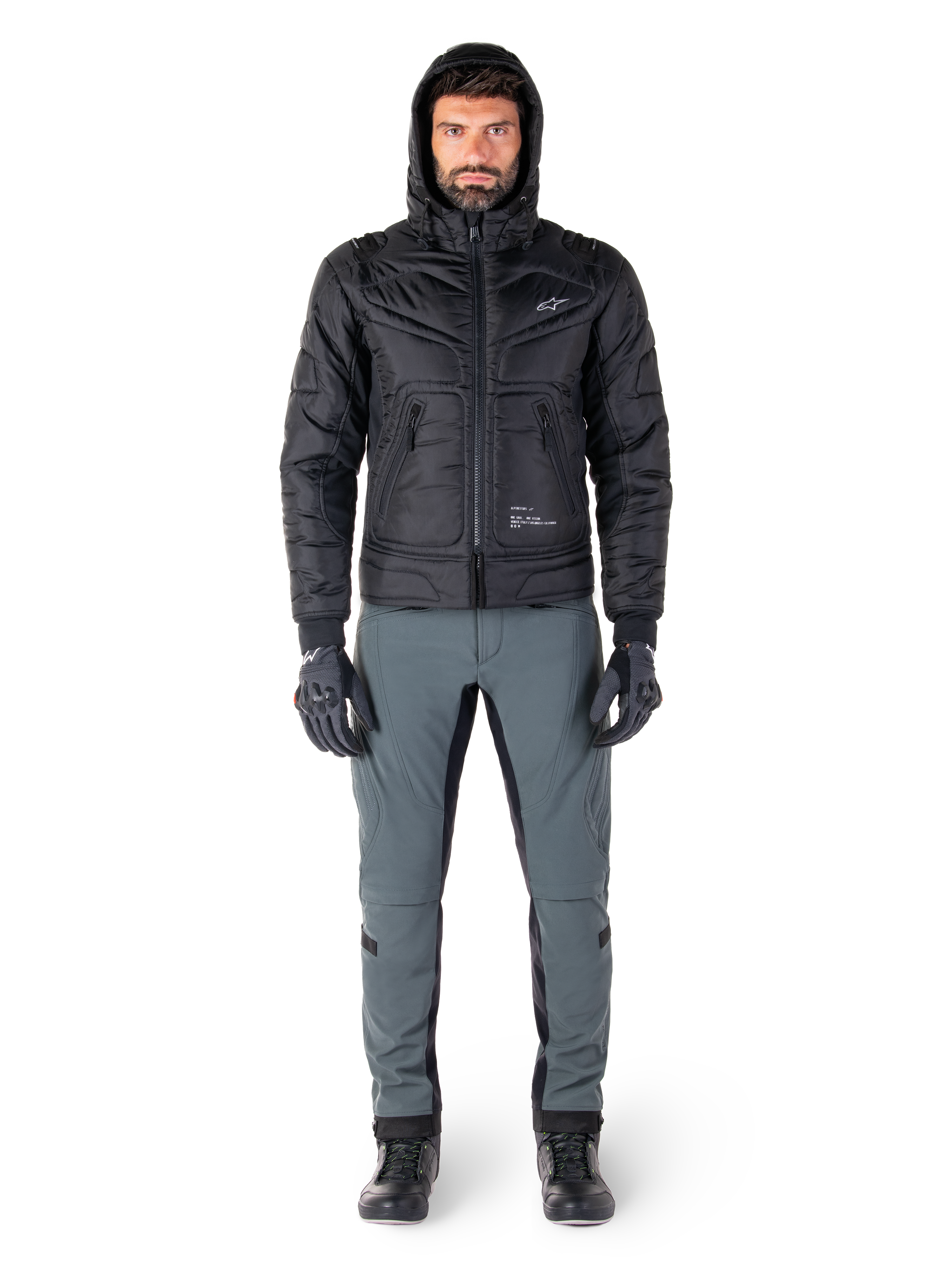 Kurtka Alpinestars Mohobbs Waterproof w kolorze czarnym, męski model w pikowanej kurtce typu bomber z kapturem, zestawiona z szarymi spodniami motocyklowymi, czarnymi rękawicami i butami, miejska współczesna odzież motocyklowa