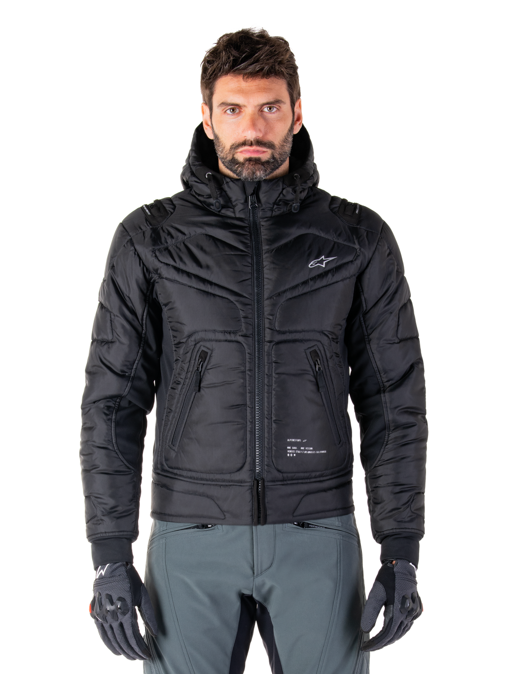 Alpinestars Mohobbs Waterproof Jacket, czarna ocieplana motocyklowa kurtka bomberka, pikowana tkanina poliestrowa z zamkiem błyskawicznym z przodu i kieszeniami na suwak, małe białe logo na piersi, noszona przez modela w szarych spodniach i rękawicach, miejski design streetwear do jazdy po mieście.