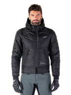 Alpinestars Mohobbs Waterproof Jacket, czarna ocieplana motocyklowa kurtka bomberka, pikowana tkanina poliestrowa z zamkiem błyskawicznym z przodu i kieszeniami na suwak, małe białe logo na piersi, noszona przez modela w szarych spodniach i rękawicach, miejski design streetwear do jazdy po mieście.