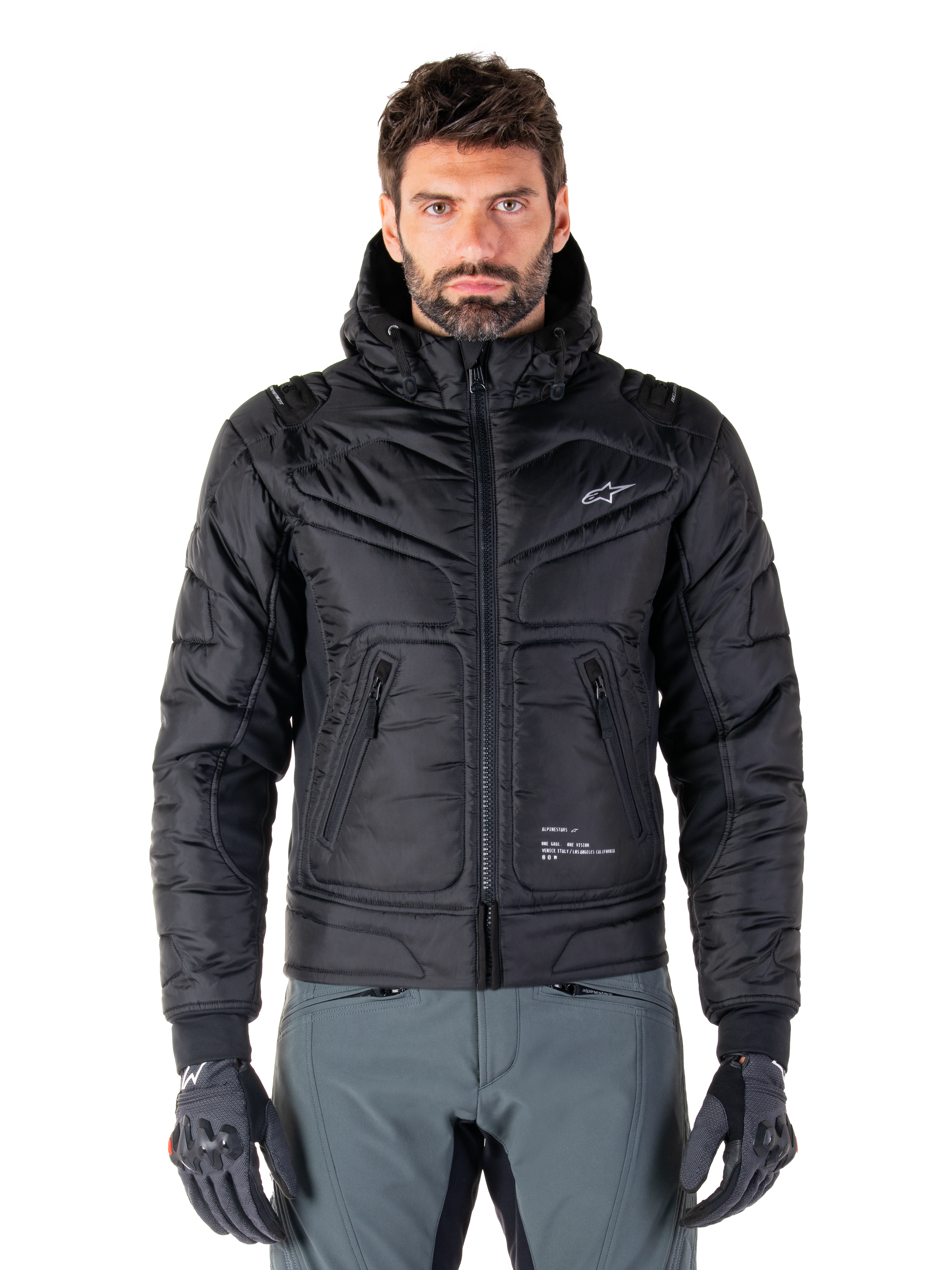 Alpinestars Mohobbs Waterproof Jacket, czarna ocieplana motocyklowa kurtka bomberka, pikowana tkanina poliestrowa z zamkiem błyskawicznym z przodu i kieszeniami na suwak, małe białe logo na piersi, noszona przez modela w szarych spodniach i rękawicach, miejski design streetwear do jazdy po mieście.