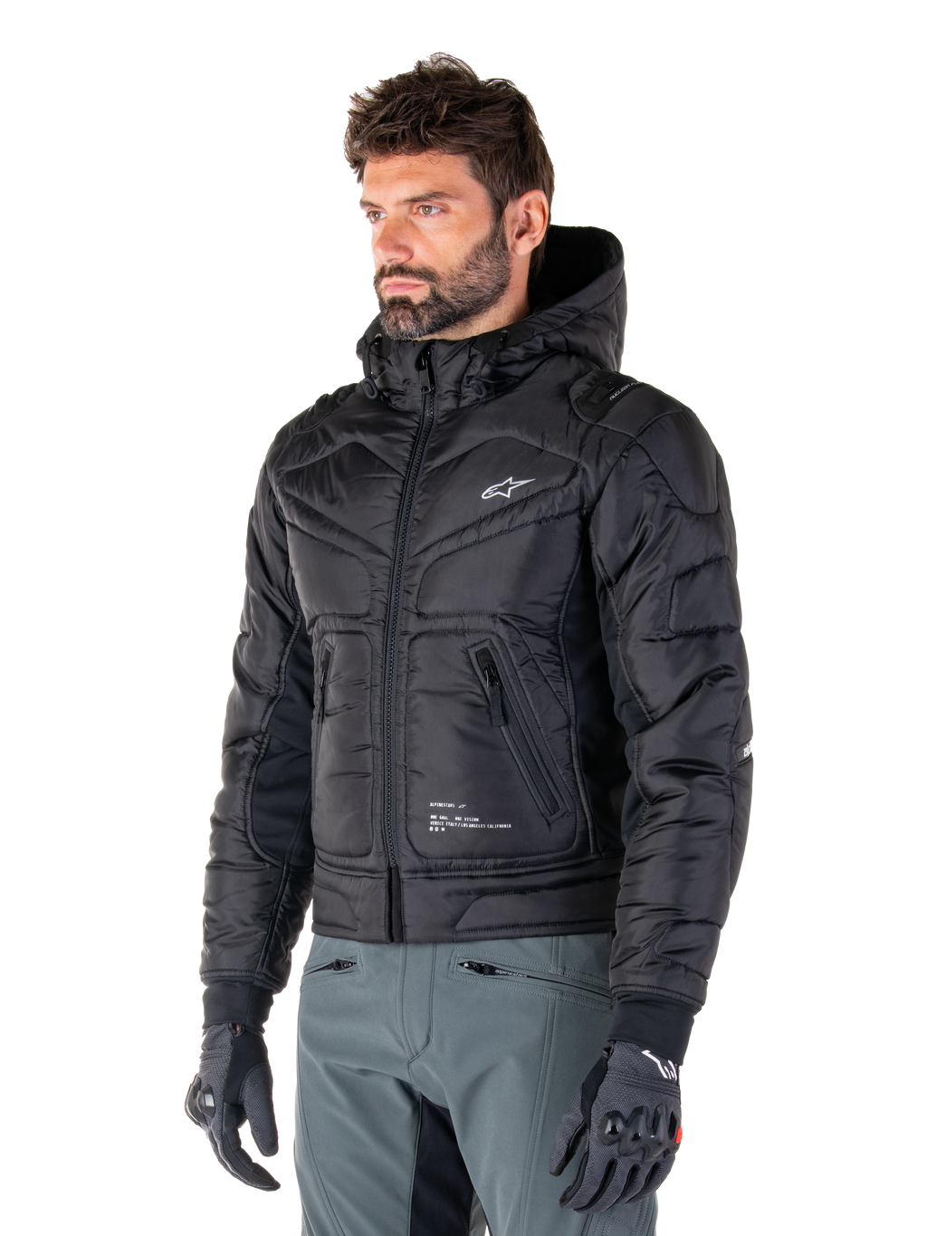Alpinestars Mohobbs Waterproof Jacket, czarna, pikowana kurtka motocyklowa typu bomber z kapturem, noszona przez mężczyznę w szarych spodniach i czarnych rękawicach, design inspirowany stylem streetwear, małe białe logo na piersi