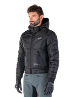 Alpinestars Mohobbs Waterproof Jacket, czarna, pikowana kurtka motocyklowa typu bomber z kapturem, noszona przez mężczyznę w szarych spodniach i czarnych rękawicach, design inspirowany stylem streetwear, małe białe logo na piersi
