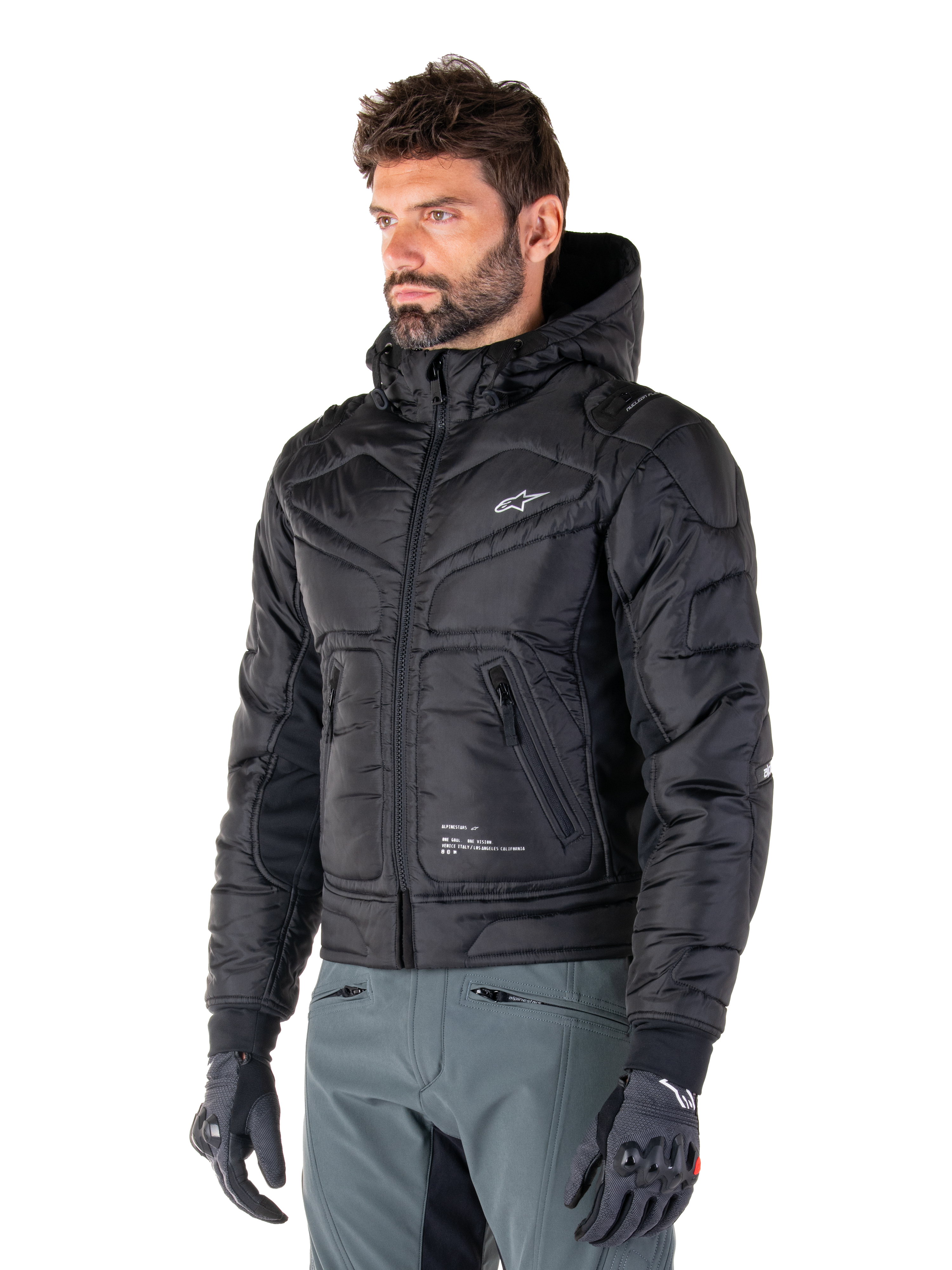 Alpinestars Mohobbs Waterproof Jacket, czarna, pikowana kurtka motocyklowa typu bomber z kapturem, noszona przez mężczyznę w szarych spodniach i czarnych rękawicach, design inspirowany stylem streetwear, małe białe logo na piersi