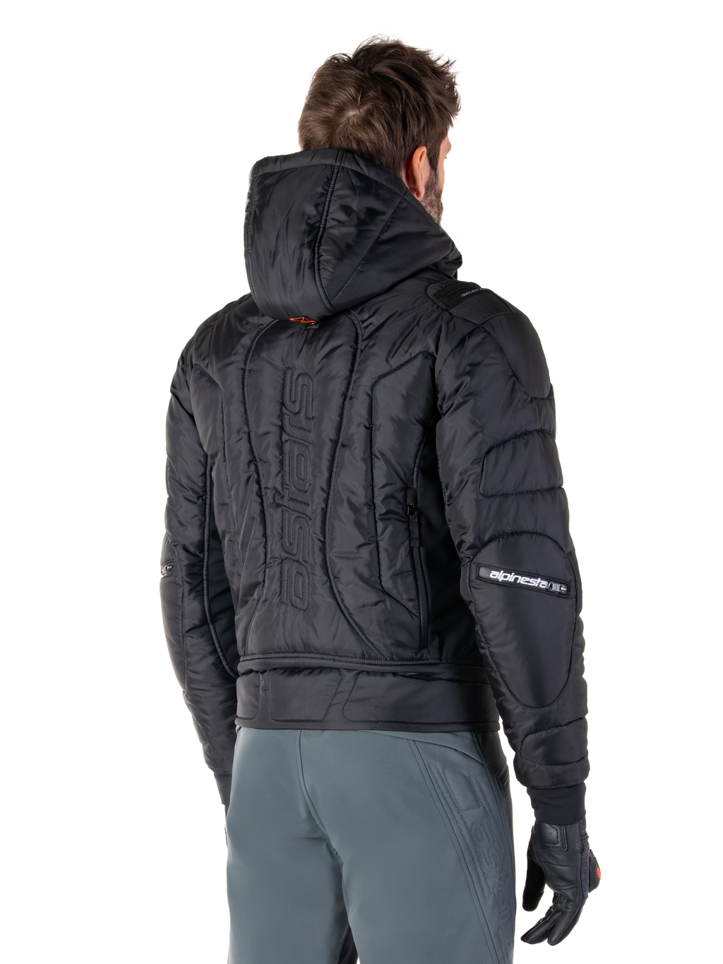 Alpinestars Mohobbs Waterproof Jacket, czarna, widok z tyłu prezentujący pikowaną, wyściełaną konstrukcję, kaptur, pionowy napis „astars” na plecach oraz branding Alpinestars na rękawach, noszona z szarymi spodniami i czarnymi rękawicami.