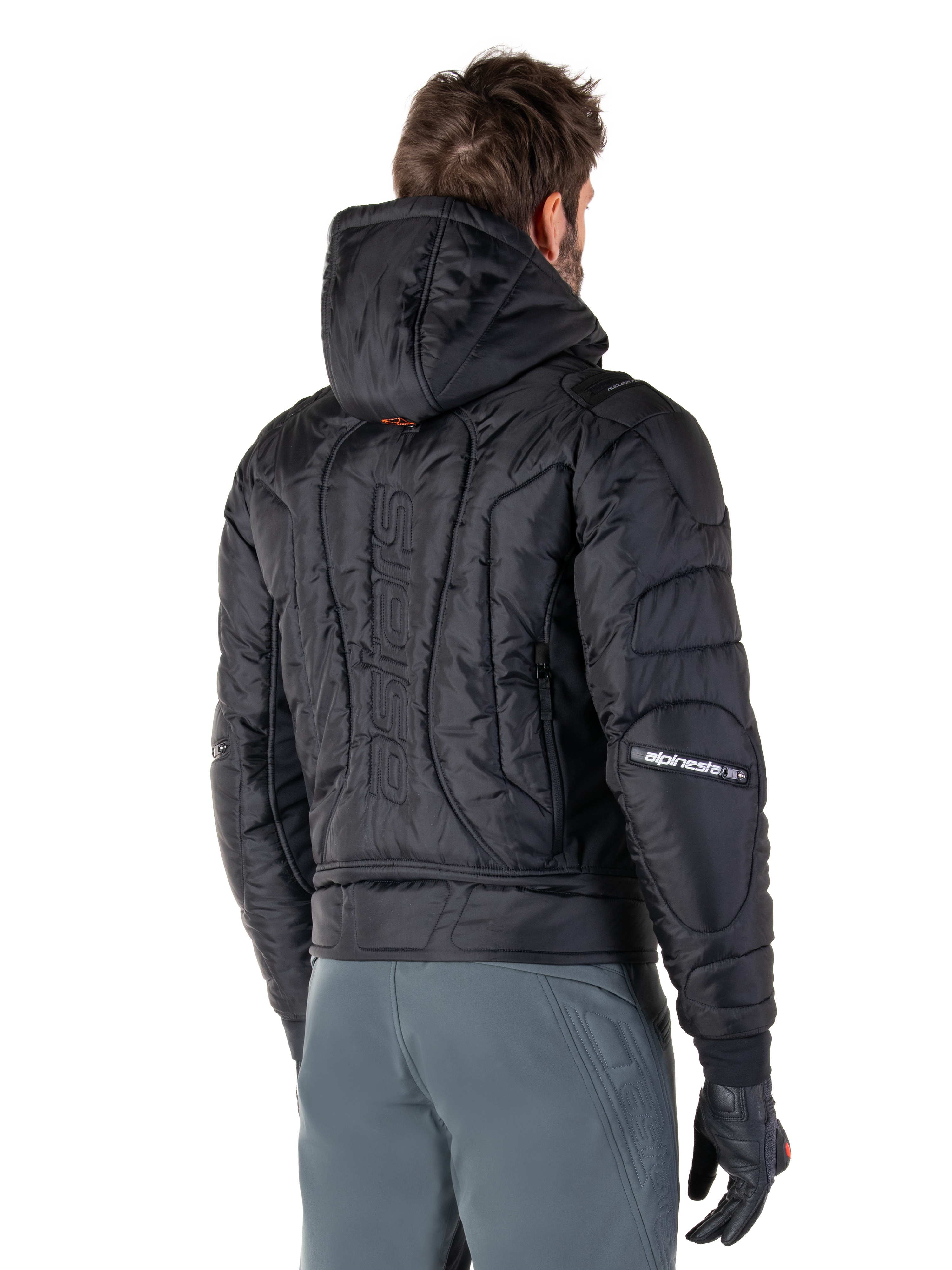 Alpinestars Mohobbs Waterproof Jacket, czarna, widok z tyłu prezentujący pikowaną, wyściełaną konstrukcję, kaptur, pionowy napis „astars” na plecach oraz branding Alpinestars na rękawach, noszona z szarymi spodniami i czarnymi rękawicami.