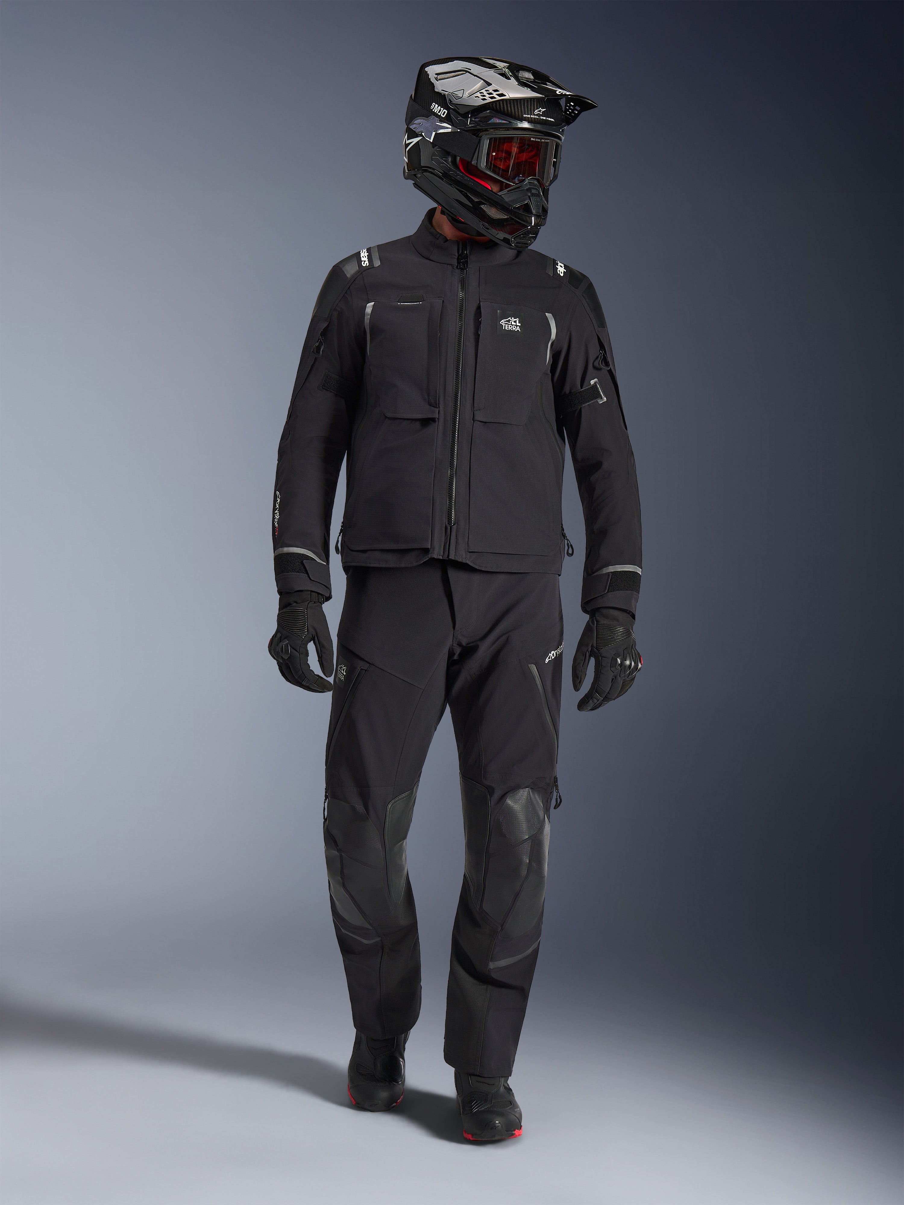 Adventure rider wearing the Alpinestars Andes Pro Drystar® XF Jacket in Black and Dark Gray, matching black adventure pants, black and white helmet with red goggles, black gloves, and boots, features large front utility pockets, reflective accents, and technical 3-layer laminated construction for all-weather touring.