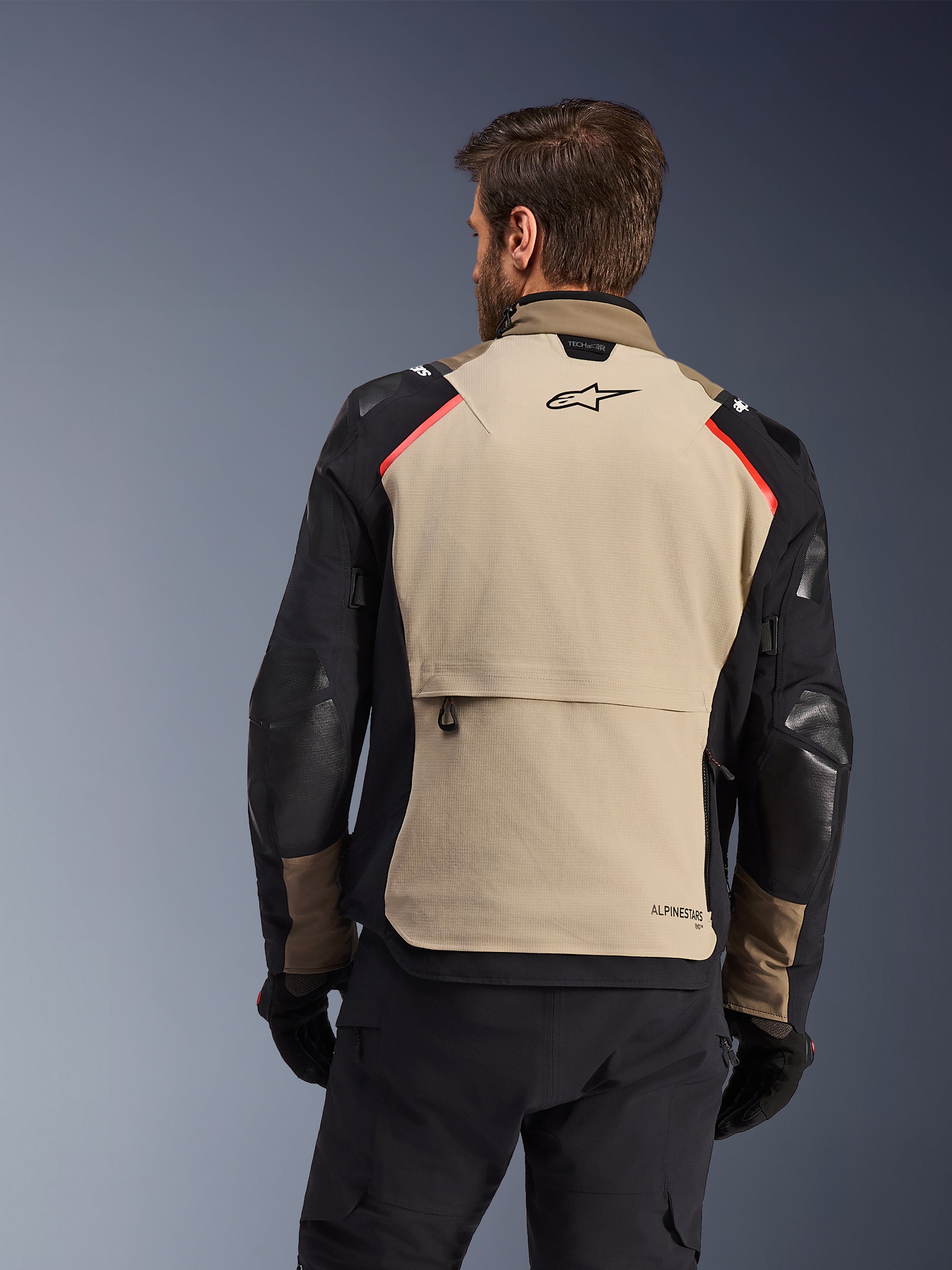 Kurtka Alpinestars Andes Pro Drystar® XF, widok z tyłu, kolor Walnut i Aluminum, z dużym beżowym panelem na plecach, czarnym logo Alpinestars i czerwonymi akcentami na ramionach, duża tylna kieszeń cargo, zestaw z czarnymi spodniami i rękawicami, odzież motocyklowa adventure touring