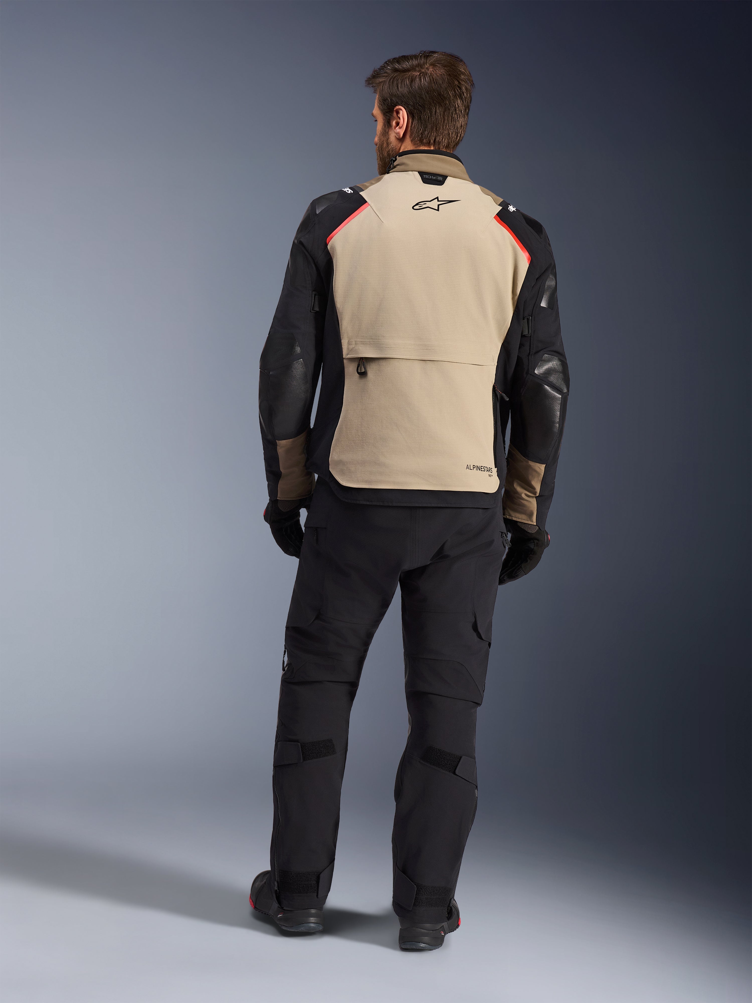 Kurtka Alpinestars Andes Pro Drystar® XF, Walnut Aluminum, widok z tyłu, motocyklowa kurtka adventure z czarnymi rękawami i czerwonymi akcentami na ramionach, noszona z czarnymi spodniami motocyklowymi i rękawicami.