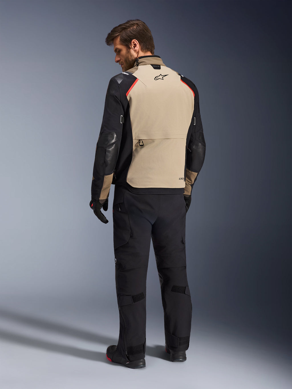 Kurtka Alpinestars Andes Pro Drystar® XF, kurtka motocyklowa, orzechowo-czarna z czerwonymi akcentami, widok z tyłu na modelu, design adventure touring z dużą tylną kieszenią, noszona z czarnymi spodniami i rękawicami motocyklowymi