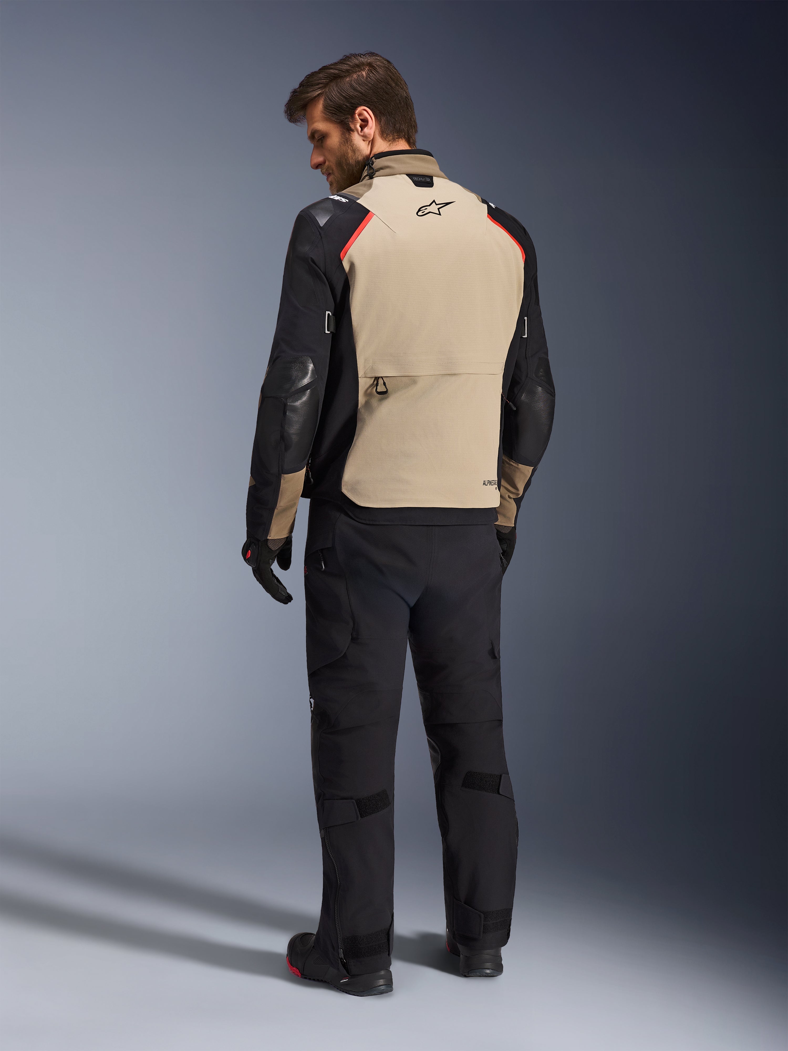 Kurtka Alpinestars Andes Pro Drystar® XF, kurtka motocyklowa, orzechowo-czarna z czerwonymi akcentami, widok z tyłu na modelu, design adventure touring z dużą tylną kieszenią, noszona z czarnymi spodniami i rękawicami motocyklowymi