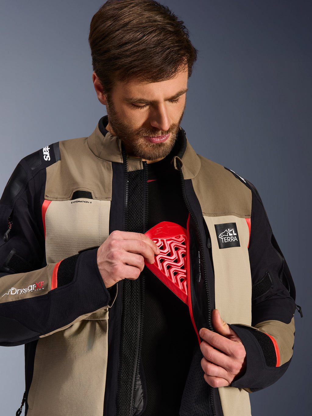 Kurtka Alpinestars Andes Pro Drystar® XF w kolorze Walnut Aluminum, model prezentujący wkładanie czerwonego ochraniacza klatki piersiowej Nucleon do wewnętrznej kieszeni, przygodowa kurtka motocyklowa z logo ALL TERRA i Drystar XF