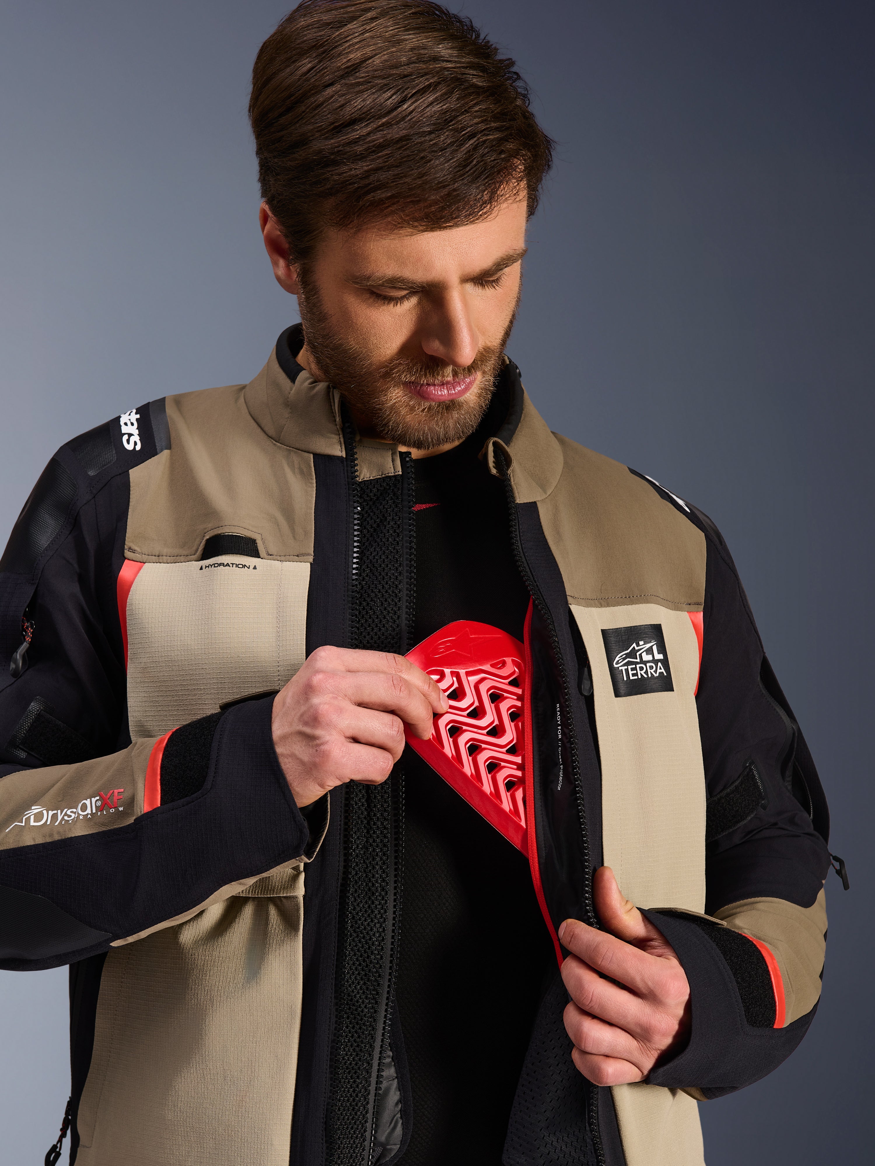 Kurtka Alpinestars Andes Pro Drystar® XF w kolorze Walnut Aluminum, model prezentujący wkładanie czerwonego ochraniacza klatki piersiowej Nucleon do wewnętrznej kieszeni, przygodowa kurtka motocyklowa z logo ALL TERRA i Drystar XF