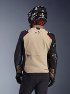 Kurtka Alpinestars Andes Pro Drystar® XF w kolorze Walnut Aluminum, widok z tyłu, piaskowy panel na plecach z dużą kieszenią cargo, czarne rękawy ze wzmocnionymi panelami, logo Alpinestars w górnej części pleców, w zestawie z czarnym kaskiem i rękawicami do jazdy adventure touring.