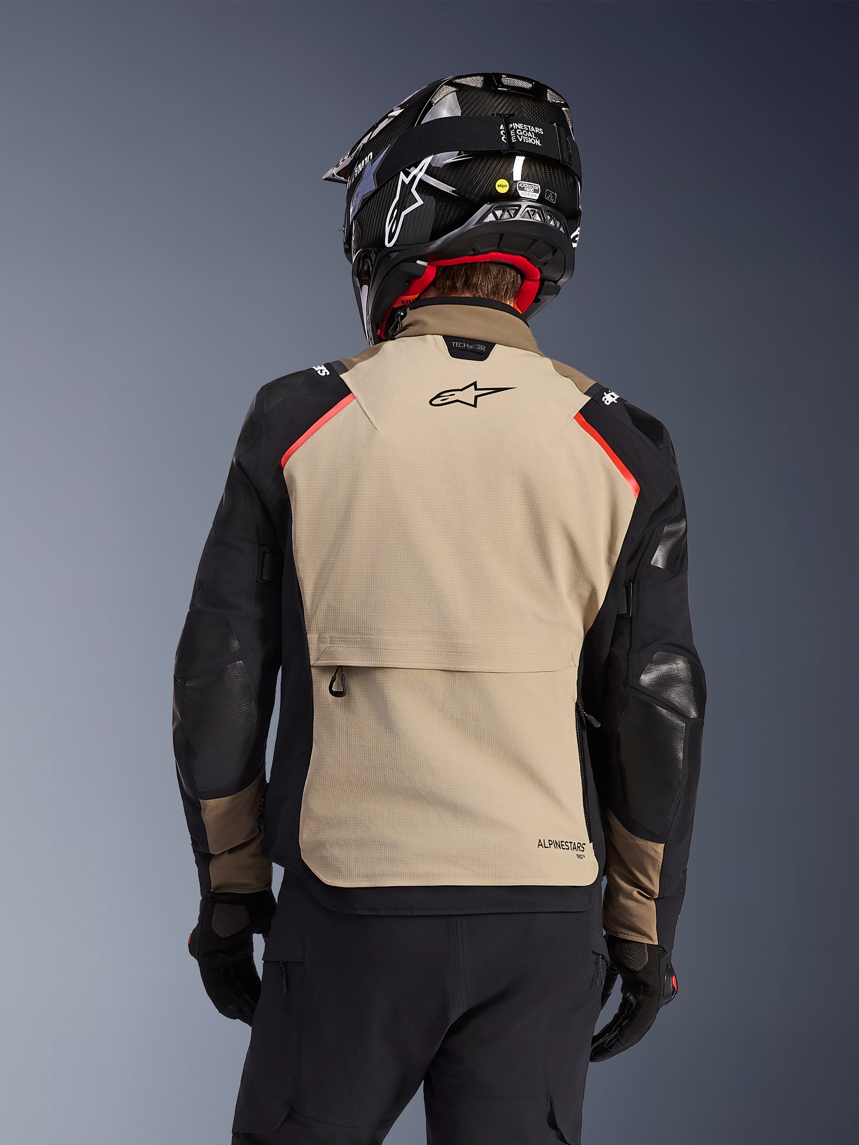 Kurtka Alpinestars Andes Pro Drystar® XF w kolorze Walnut Aluminum, widok z tyłu, piaskowy panel na plecach z dużą kieszenią cargo, czarne rękawy ze wzmocnionymi panelami, logo Alpinestars w górnej części pleców, w zestawie z czarnym kaskiem i rękawicami do jazdy adventure touring.