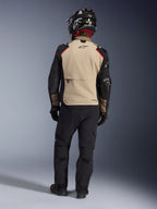 Motocyklista widoczny od tyłu w kurtce Alpinestars Andes Pro Drystar® XF w kolorze Walnut Aluminum, beżowy panel pleców z czarnymi rękawami i czerwonymi akcentami, czarne techniczne spodnie adventure i kask motocyklowy, odzież touringowa premium na każdą pogodę.