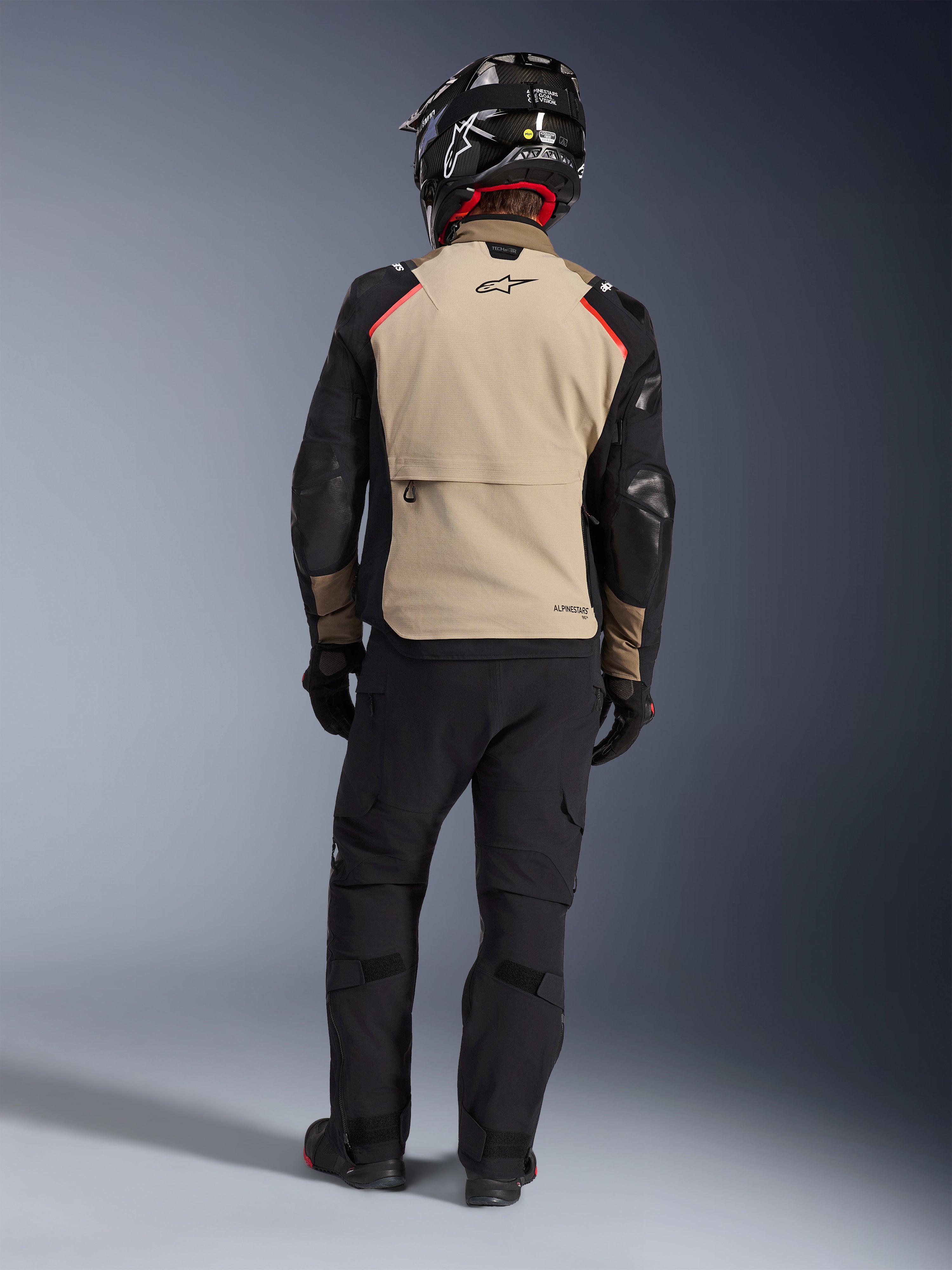 Motocyklista widoczny od tyłu w kurtce Alpinestars Andes Pro Drystar® XF w kolorze Walnut Aluminum, beżowy panel pleców z czarnymi rękawami i czerwonymi akcentami, czarne techniczne spodnie adventure i kask motocyklowy, odzież touringowa premium na każdą pogodę.