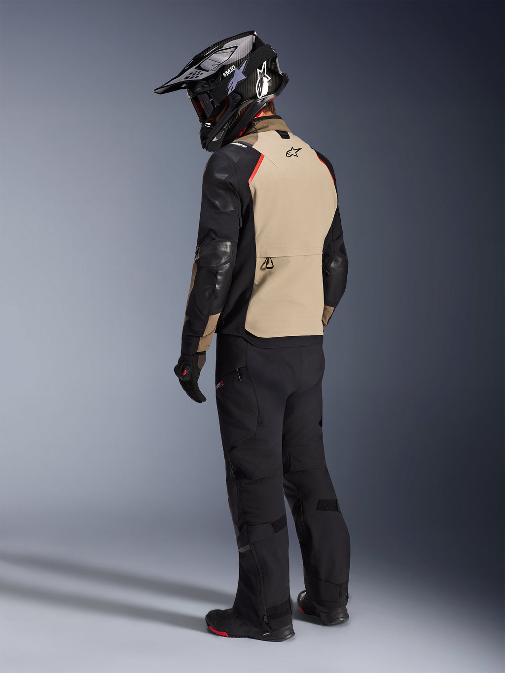 Motocyklista widziany od tyłu w kurtce Alpinestars Andes Pro Drystar® XF w kolorze Walnut Aluminum, beżowy panel tylny, czarne rękawy i czerwone akcenty, czarny kask adventure, czarne spodnie motocyklowe i buty, profesjonalna odzież adventure touring