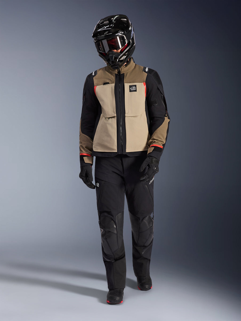 Kurtka Alpinestars Andes Pro Drystar® XF w kolorze Walnut Aluminum, zestawiona z czarnymi spodniami adventure, czarnymi rękawicami i czarnym kaskiem z goglami, z czerwonymi akcentami i brandingiem All Terra, profesjonalny zestaw adventure touring