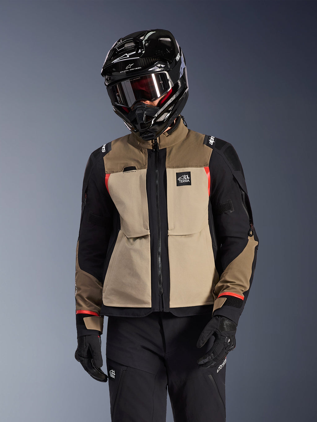 Kurtka Alpinestars Andes Pro Drystar® XF, motocyklowa kurtka adventure touring, beżowo-brązowa (Walnut Aluminum) z czarnymi panelami i czerwonymi akcentami, noszona z czarnym kaskiem i goglami Alpinestars, techniczna odzież motocyklowa