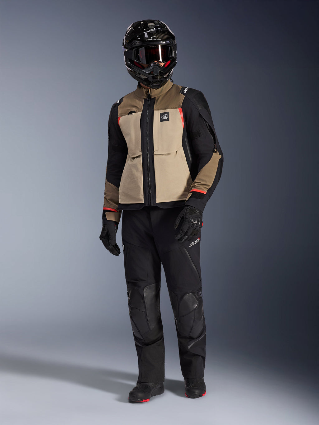 Kurtka Alpinestars Andes Pro Drystar® XF w kolorze Walnut Aluminum, motocyklowa kurtka adventure noszona z pasującymi czarnymi spodniami, czarnym kaskiem adventure z goglami i czarnymi butami, z beżowymi i czarnymi panelami oraz czerwonymi akcentami.